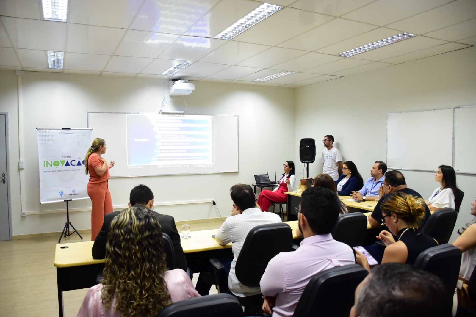Imagem colorida mostra uma mulher apresentando um projeto enquanto várias pessoas numa sala assistem.