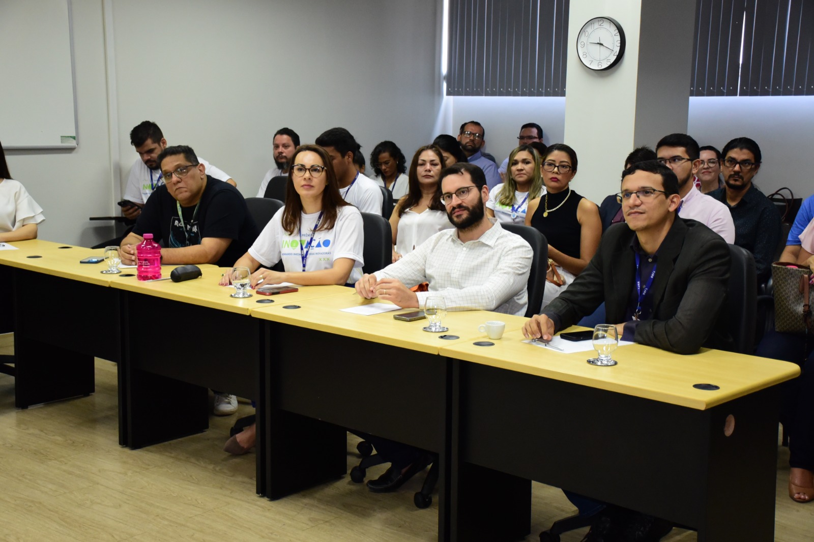 Imagem colorida mostra pessoas atrás de uma mesa de madeira enquanto assistem uma apresentação.