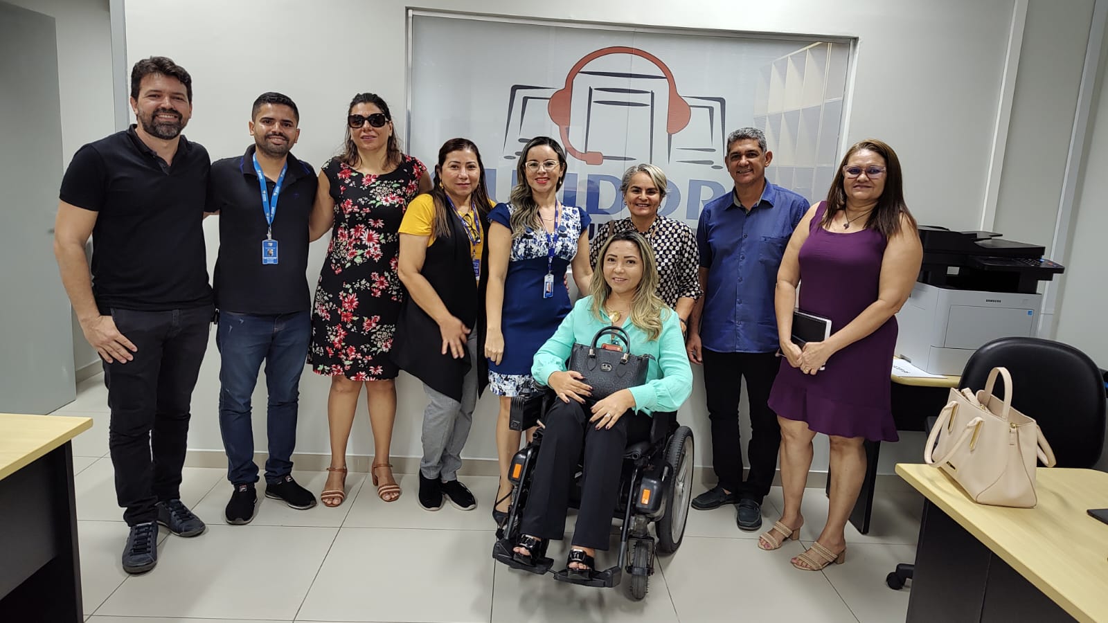 Imagem colorida mostra um grupo de nove pessoas servidoras do TJRR, reunidas posando para fotografia com o símbolo da Ouvidoria-Geral ao fundo