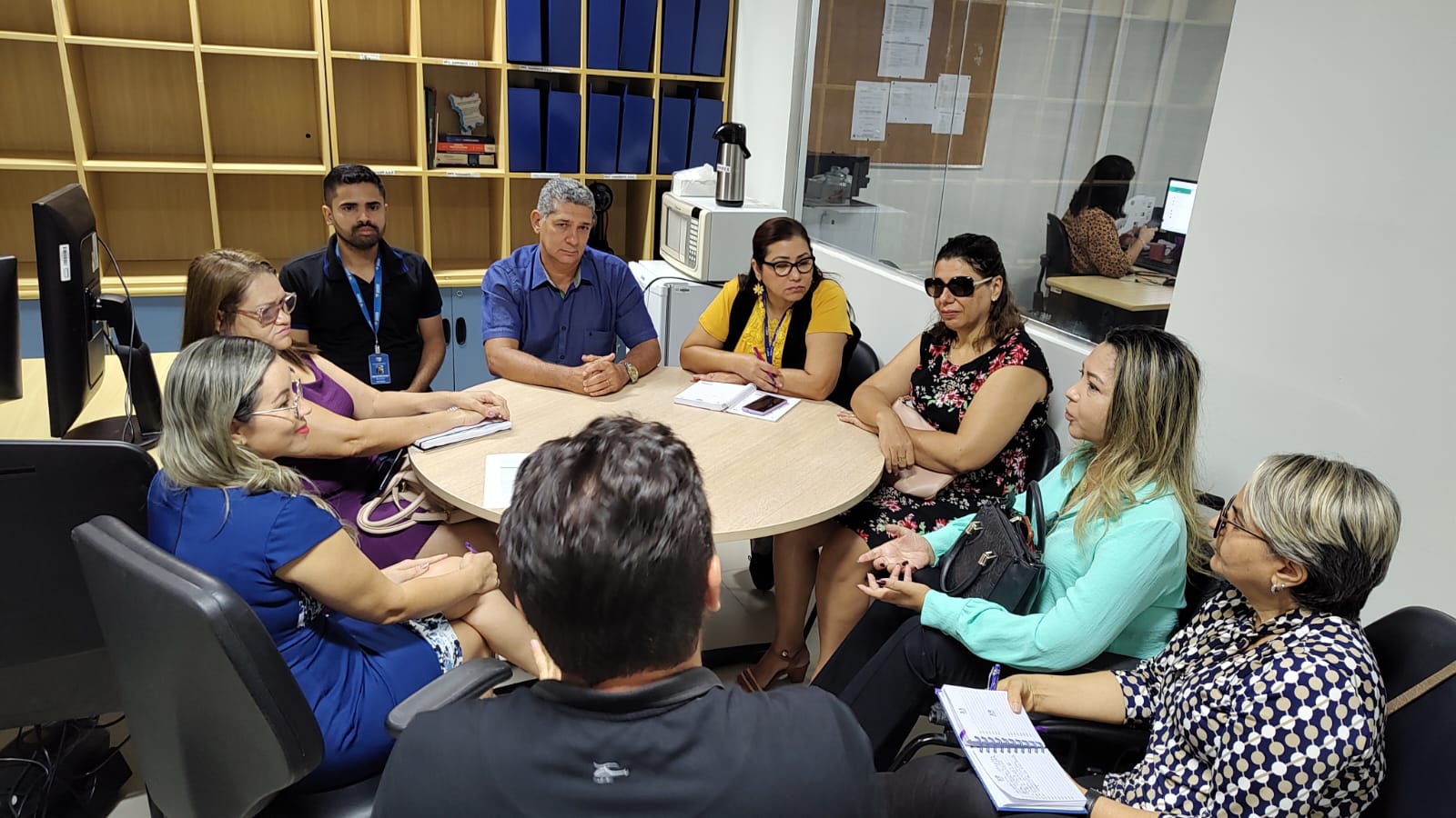 Imagem colorida mostra um grupo de nove pessoas servidoras do TJRR, reunidas ao redor de uma mesa redonda, durante reunião. 