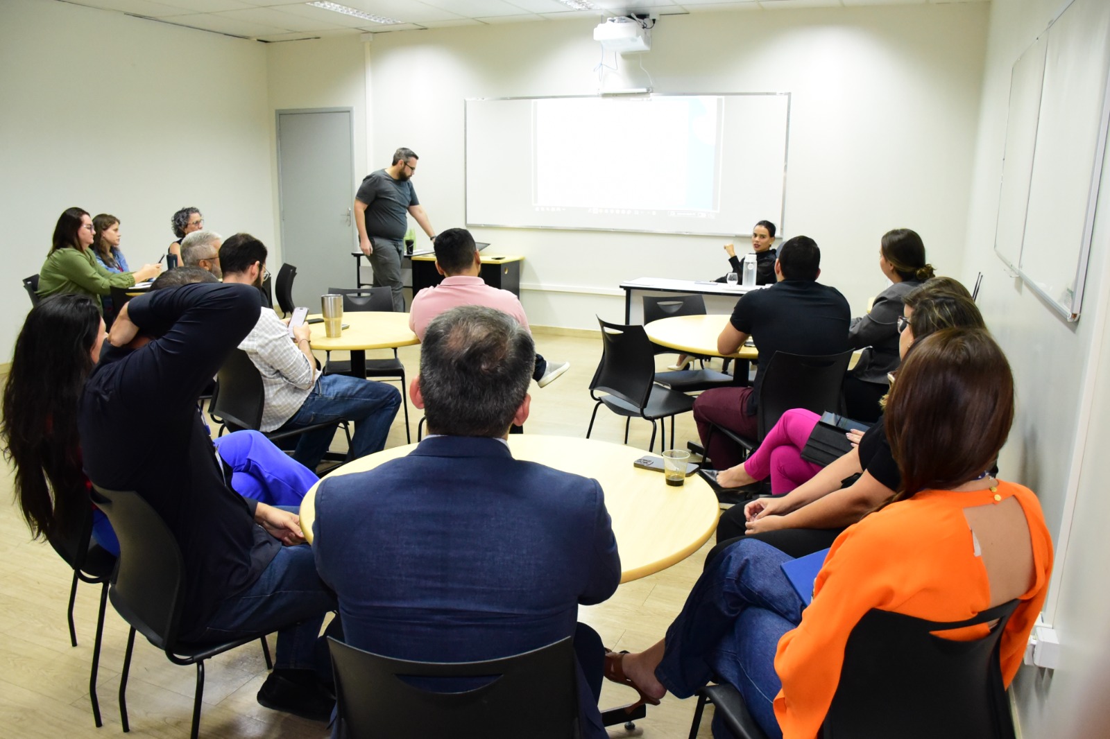foto colorida em sala com varias pessoas sentadas ao redor de mesas redondas e em direção a lousa branca