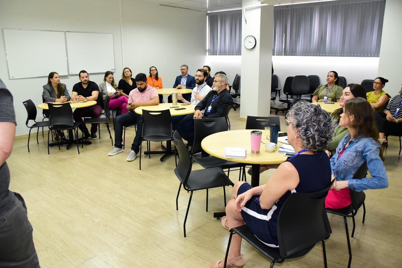 foto colorida em sala com varias pessoas sentadas ao redor de mesas redondas e em direção a lousa branca