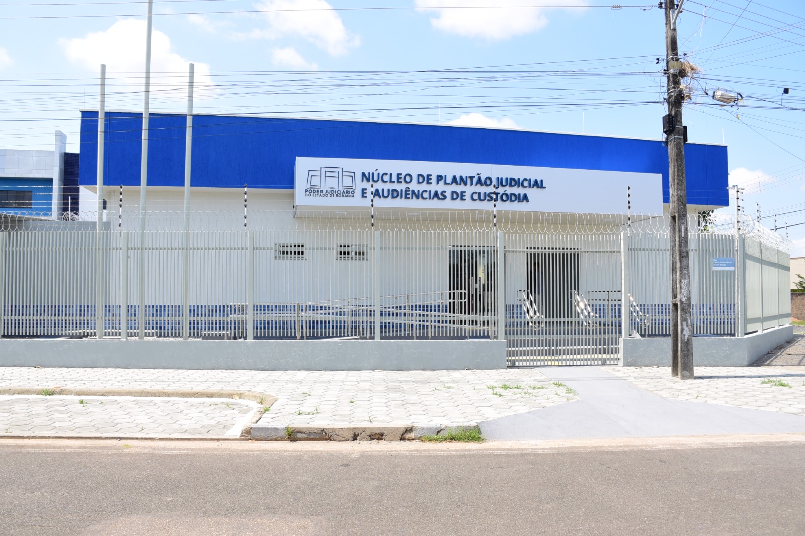 magem colorida da fachada do NUPAC feita de dia. A fachada do prédio é branca e azul, e está escrito “ Núcleo de Plantão Judicial e Audiências de Custódia” ao lado da logo do Poder Judiciário de Roraima.