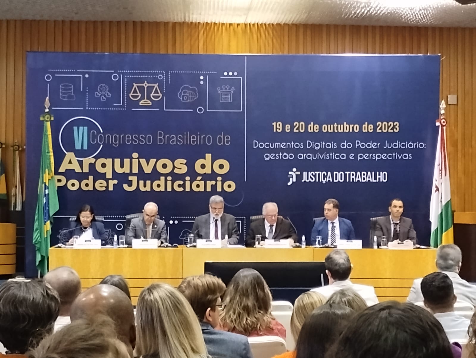 Imagem colorida mostra os seis palestrantes na bancada do VI Congresso Brasileiro de Arquivos do Poder Judiciário, sentados um ao lado do outro. Abaixo a frase “GESTÃO DOCUMENTAL - Congresso Brasileiro de Arquivos do Poder Judiciário Promove Intercâmbio de Conhecimento.” 