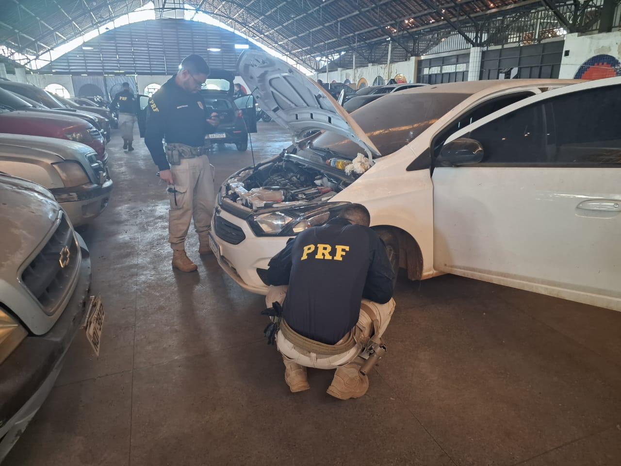 foto colorida do galpão com dois policiais da PRF inspecionando um carro branco 