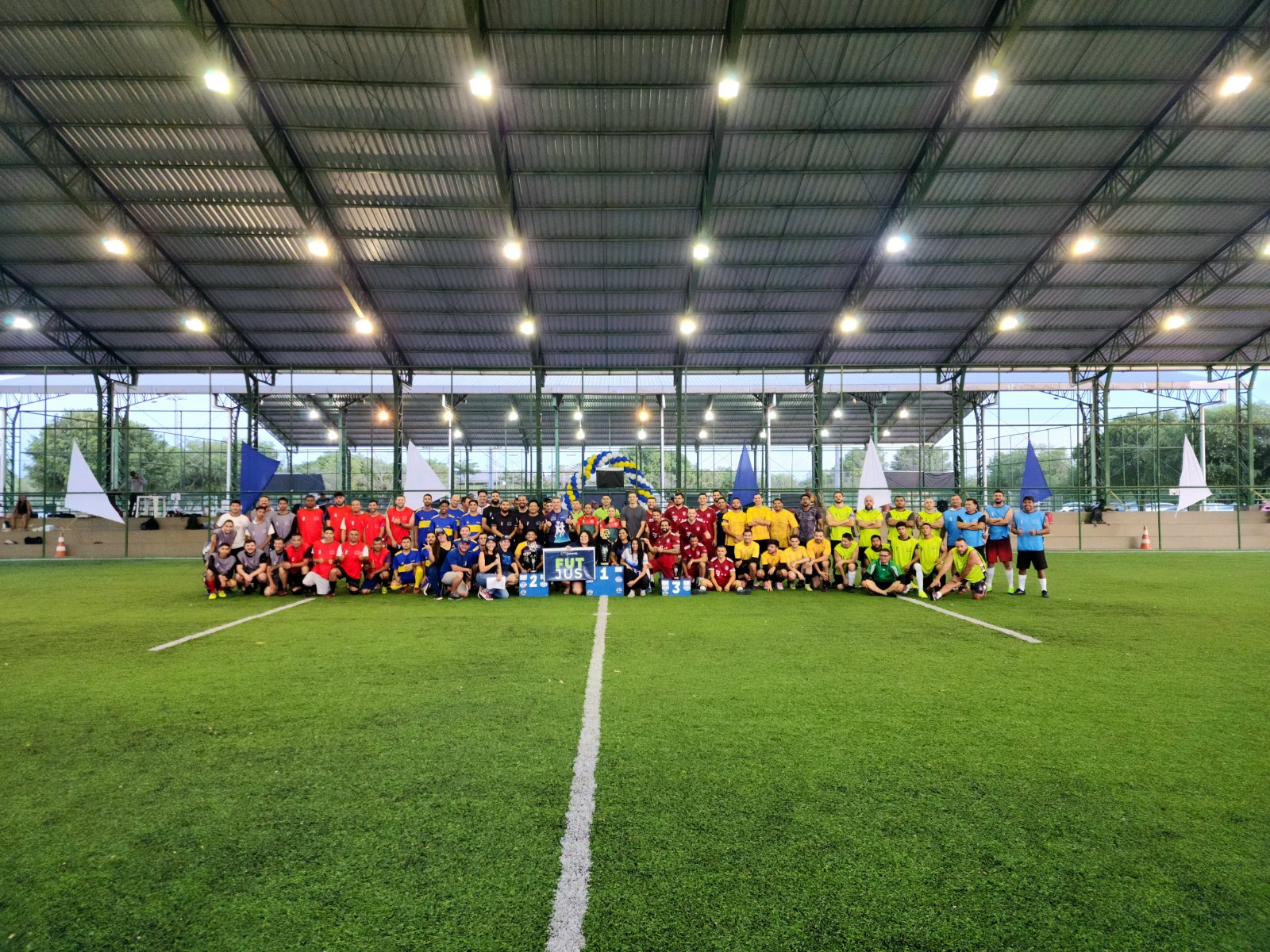 Imagem colorida mostra oito times de futebol society posando para fotografia no campo de futebol com servidores da Secretaria de Qualidade de Vida (SQV/TJRR) e o um trio de juízes de futebol, durante a 4ª edição do FutJus, que ocorreu na Vila Olímpica Roberto Marinho.