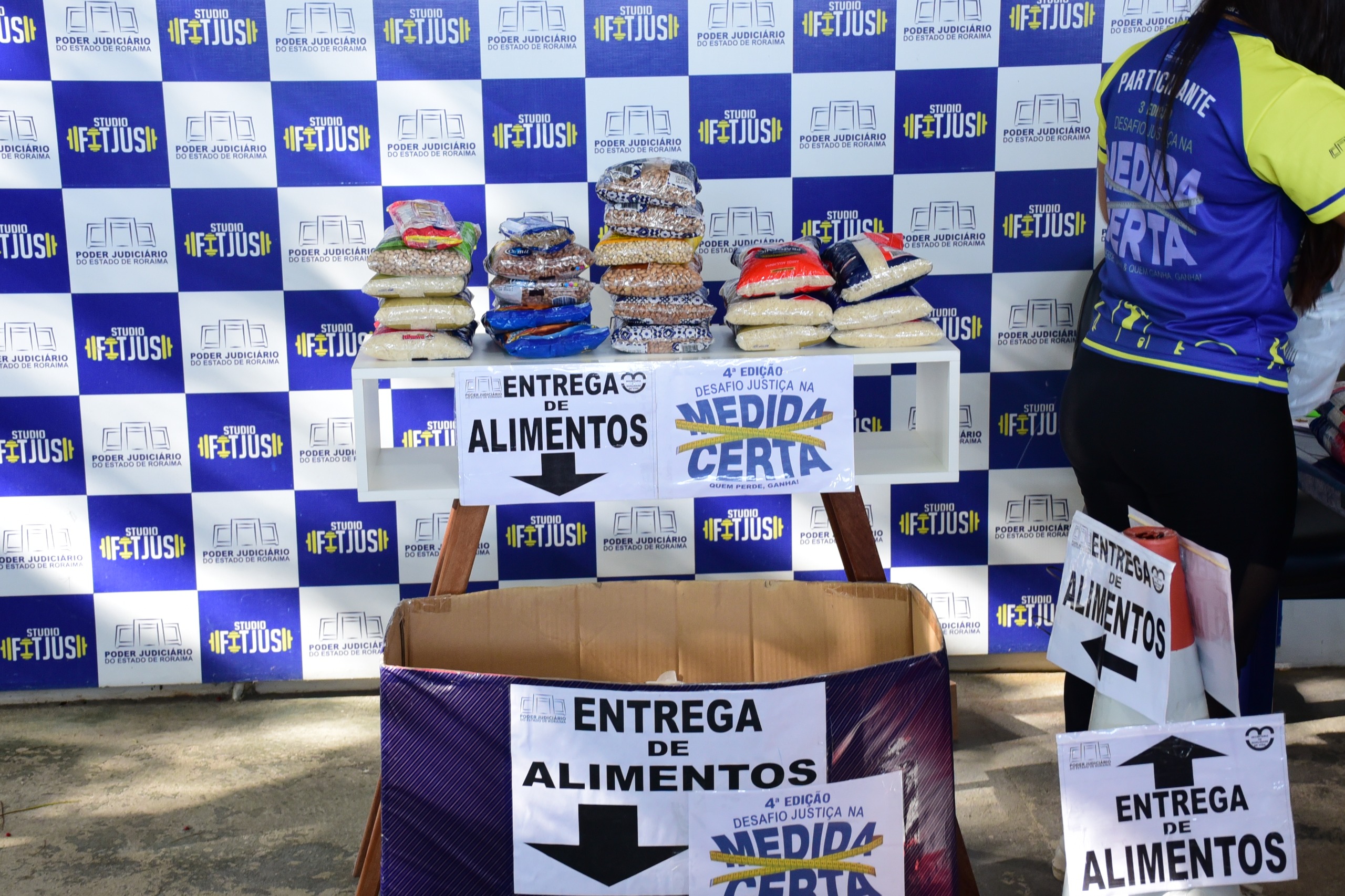 foto colorida de uma mesa com alimentos em cima e com papéis colados escrito “entrega de alimentos” e 4° edição desafio justiça na medida certa” e abaixo uma caixa de papelão azul para ser depositado os alimentos. Ao fundo é possível ver o backdrop com a logo do TJRR e do “Studio Fitjus”. 