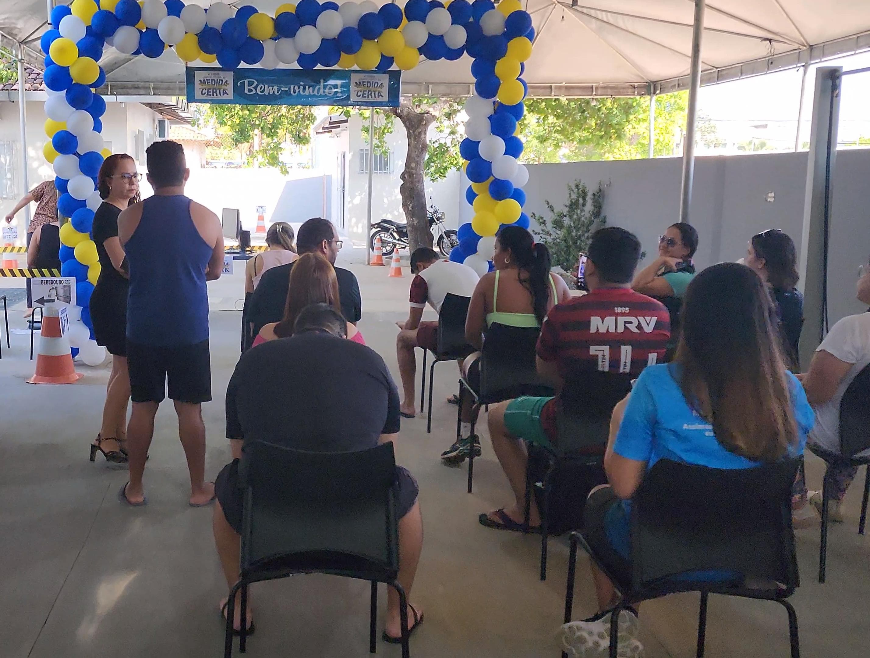  foto colorida de servidores do TJRR sentados em cadeiras enquanto aguardam serem atendidos.Na entrada há um portal com balões amarelo, branco e azul e com uma faixa “Bem vindo”. 