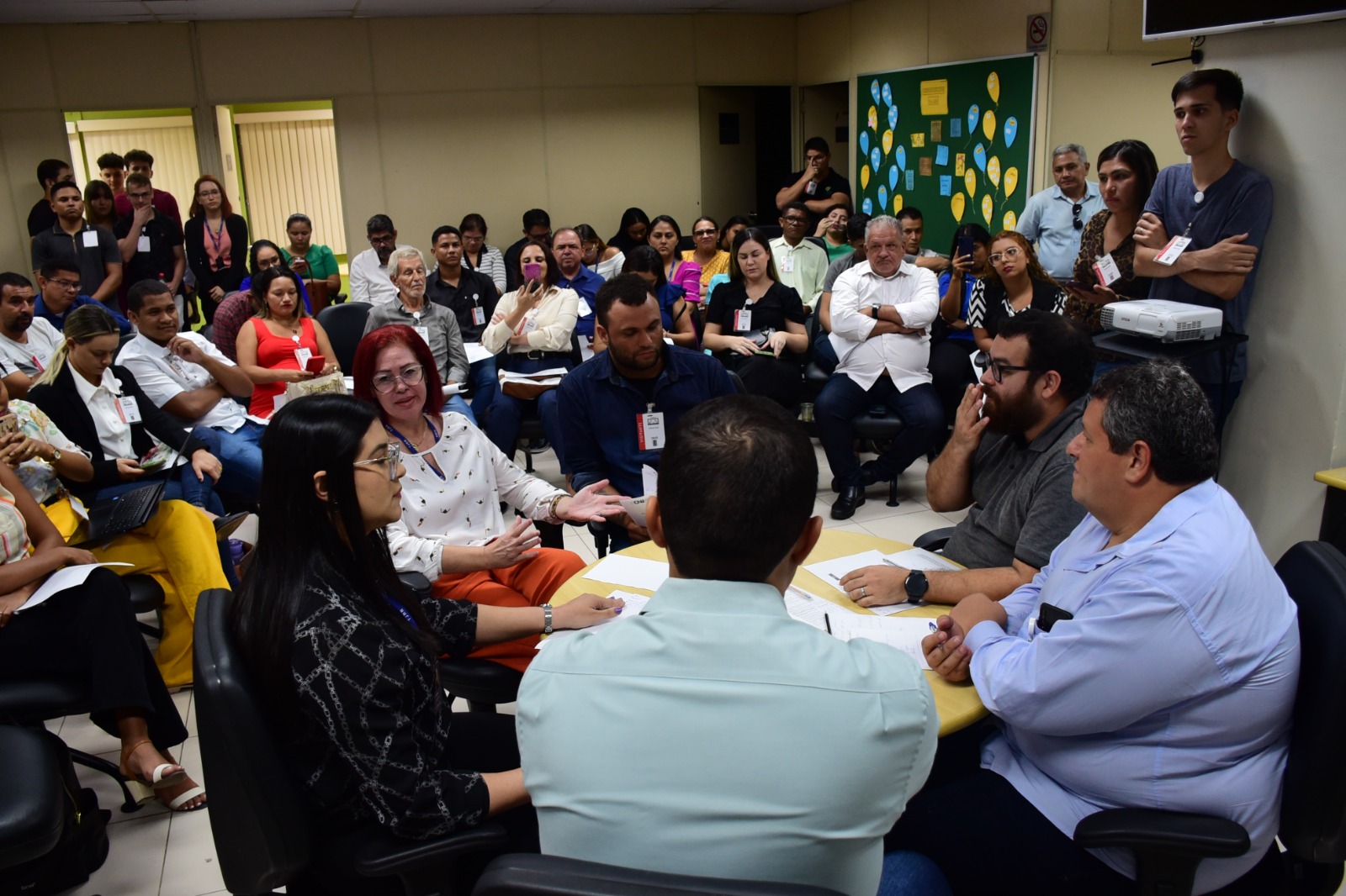 Imagem colorida de várias pessoas reunidas em círculo em uma sala ao redor de um homem que está em pé falando.