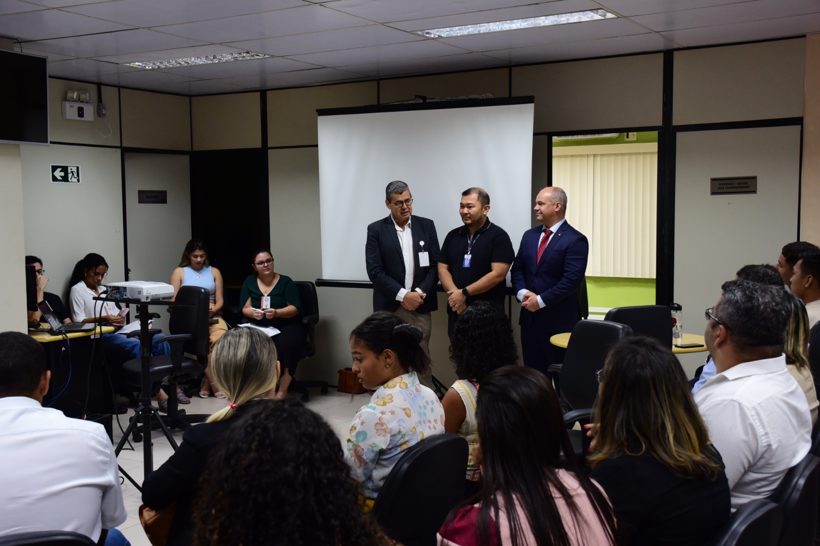 Imagem colorida de três homens em pé em uma sala, com pessoas sentadas em cadeiras ao seu redor