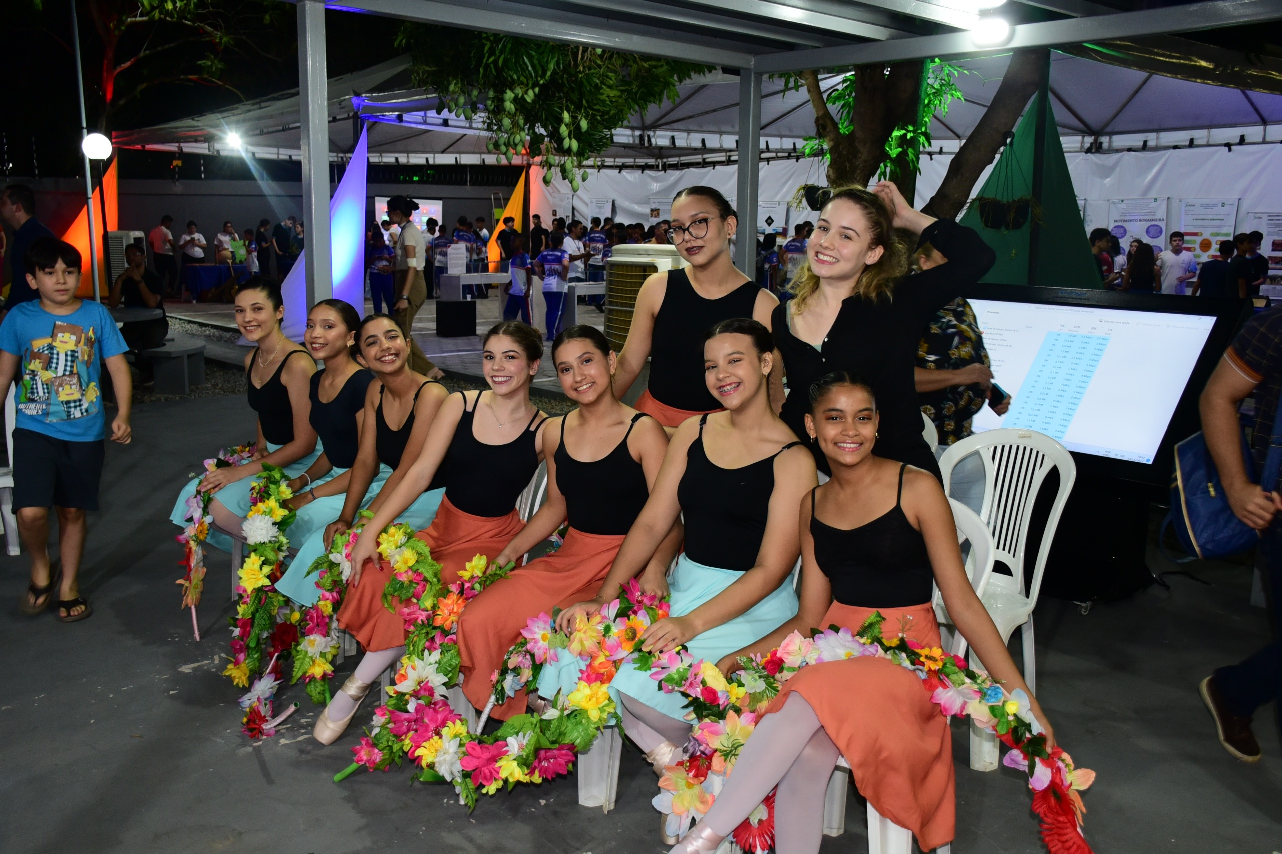 foto colorida da turma de ballet com sete sentadas e duas em pé atrás e posando para a fotografia. Elas estão segurando um arco de flores.