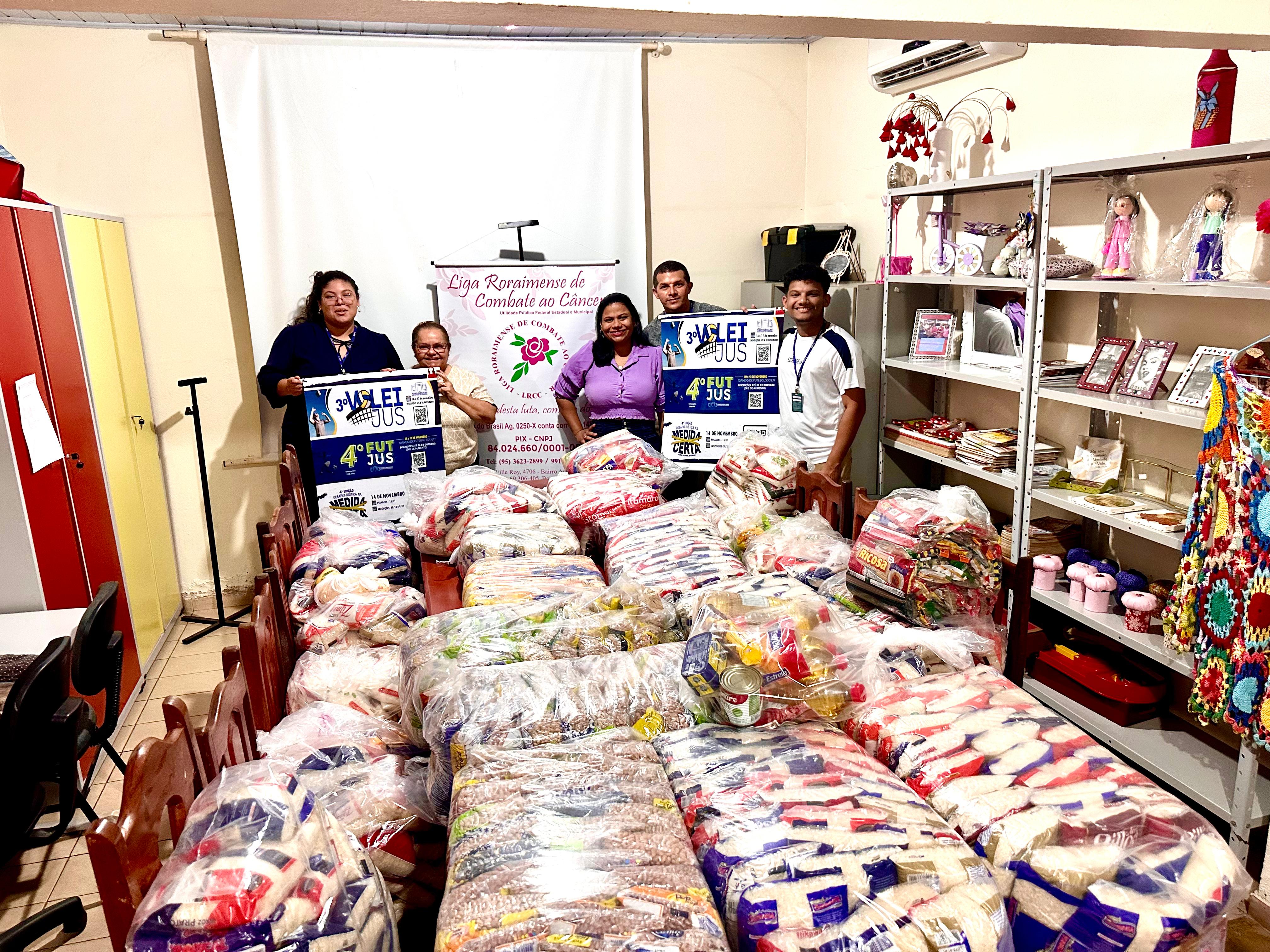 FOTO COLORIDA DE UMA SALA DA LIGA RORAIMENSE DO COMBATE AO CANCER COM VARIOS FARDOS DE ALIMENTOS. NA FOTO HÁ 5 PESSOAS POUSANDO PARA FOTO SEGURANDO O BANNER DO VOLEIJUS