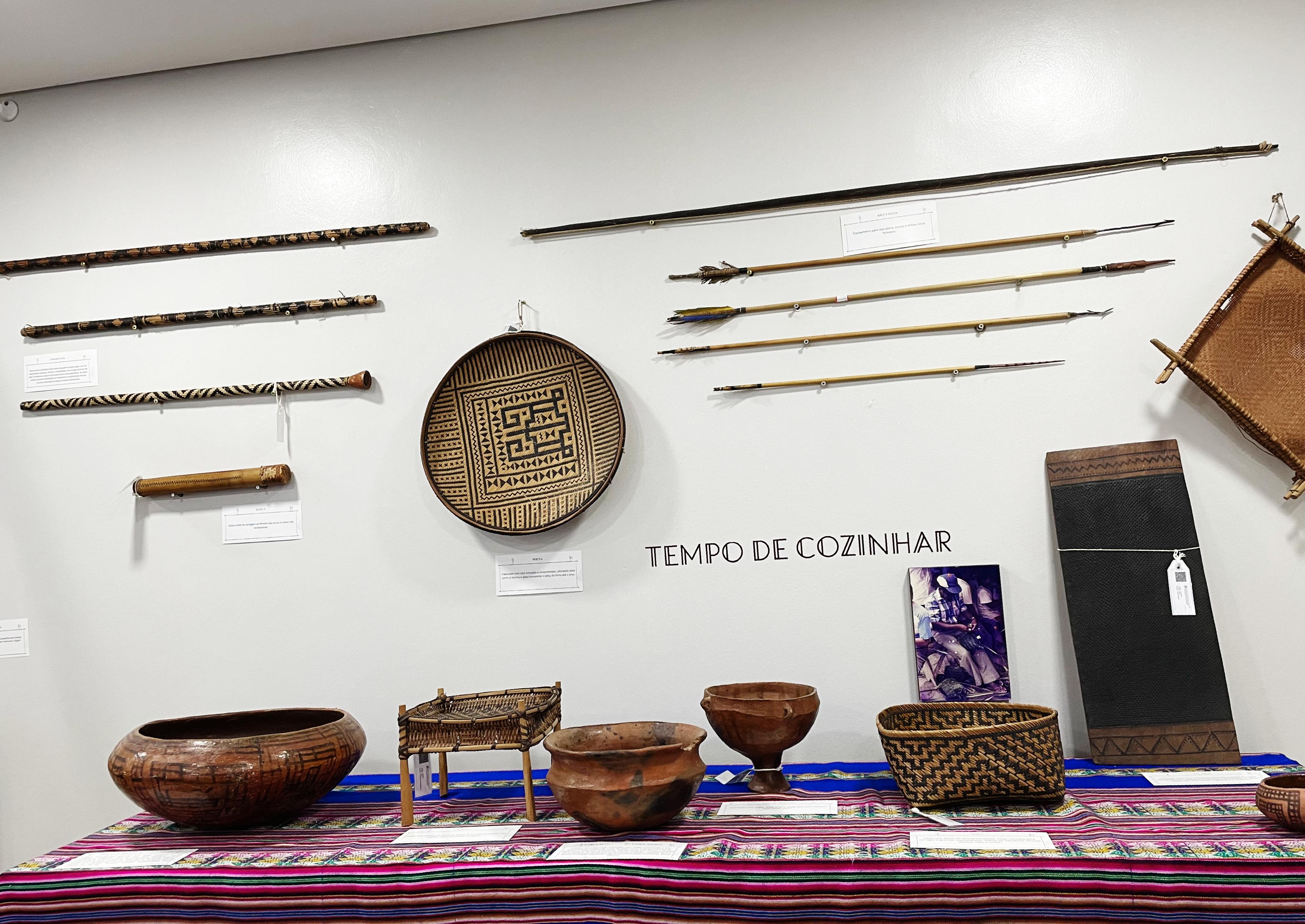 Foto colorida de cestas, flechas e panelas de barro indígenas que estão expostos em uma mesa e acima, na parede, está adesivado “tempo de cozinhar”