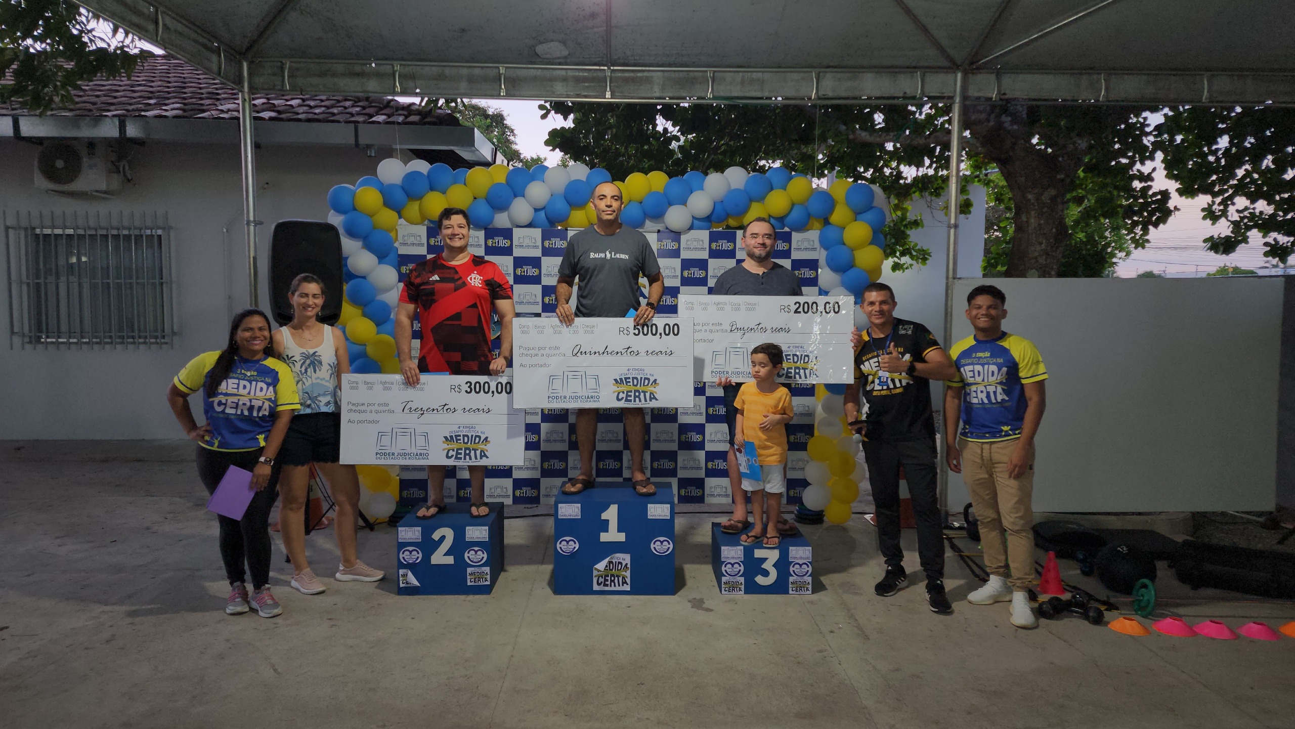 foto colorida de ganhadores homens no pódio de 1°, 2°, 3° lugar segurando as placas com valores da premiação ao lado delas estão os servidores da Secretaria de Qualidade de Vida.