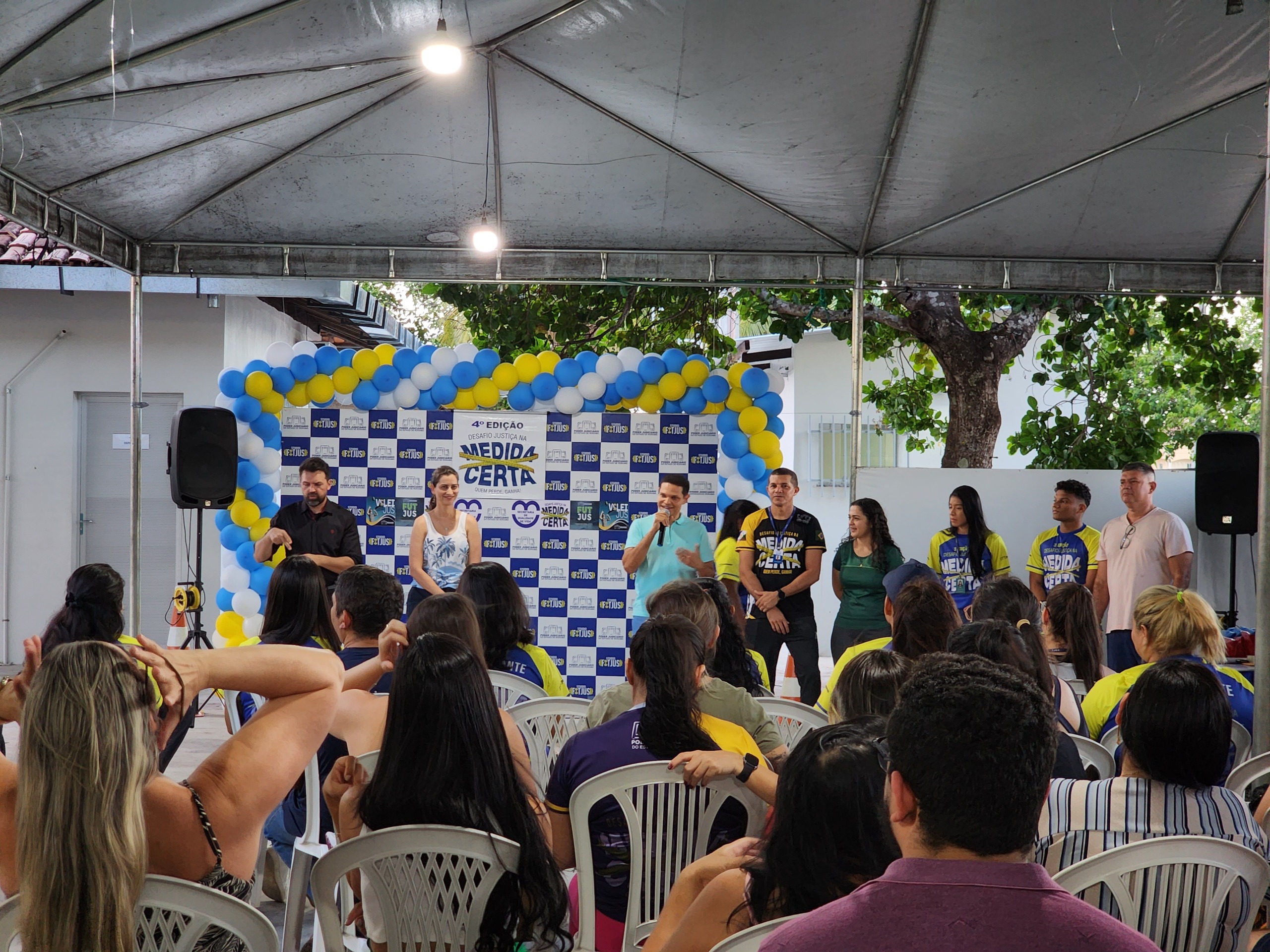 foto colorida da cerimônia de premiação ao lado esquerdo há um intérprete de libras e várias pessoas sentadas em cadeiras de plastico brancas. Abaixo está o texto “MEDIDA CERTA - Encerramento da 4ª edição do desafio conta com entrega de premiação e sorteio de brindes’’