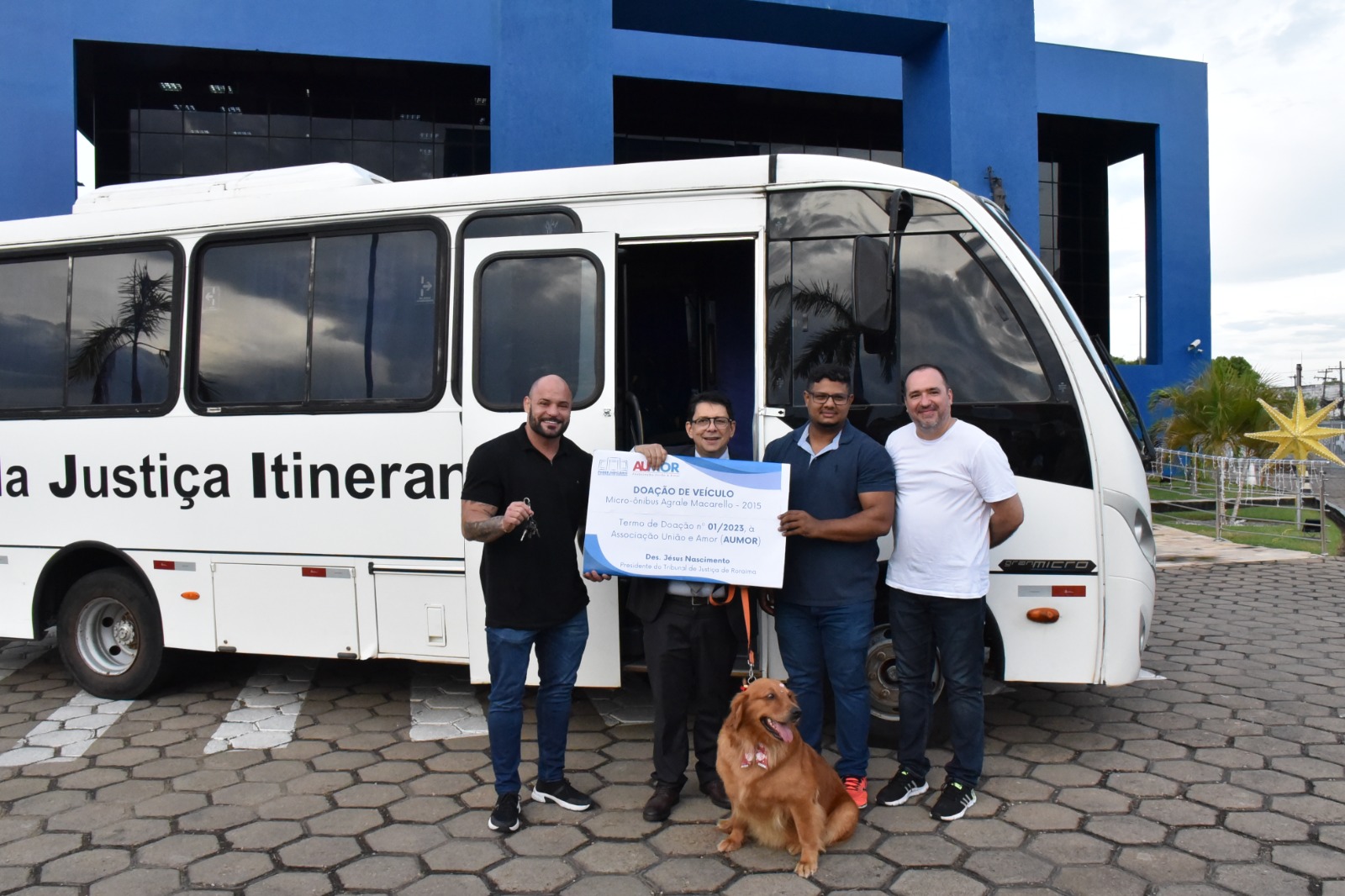 foto colorida do Presidente do Tribunal do TJRR, desembargador Jesus Nascimento junto com o deputao Marcinho Belota segurando a chave do onibus juntamente com uma placa de doação. Na foto ainda há mais um homem e uma cachorrinha 