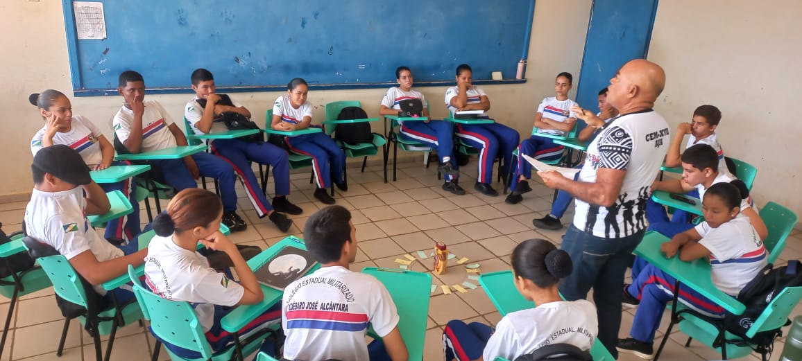 Foto colorida mostrando alunos reunidos em círculos, enquanto um professor está no meio deles, durante uma das rodas de conversas realizadas pelo Programa. 