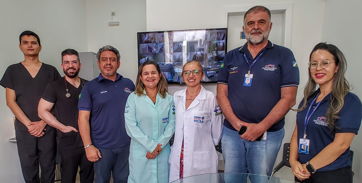 Foto colorida de servidores do TJRR e de profissionais da saúde reunidos em frente a um monitor com imagens de câmeras de segurança