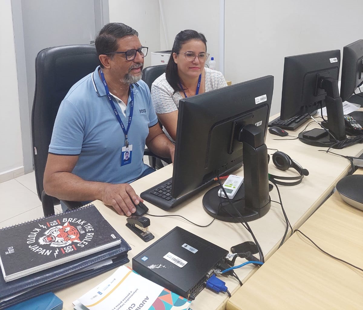 foto colorida de um servidor e uma servidora do TJRR sentado em uma estação de trabalho com três computadores na mesa