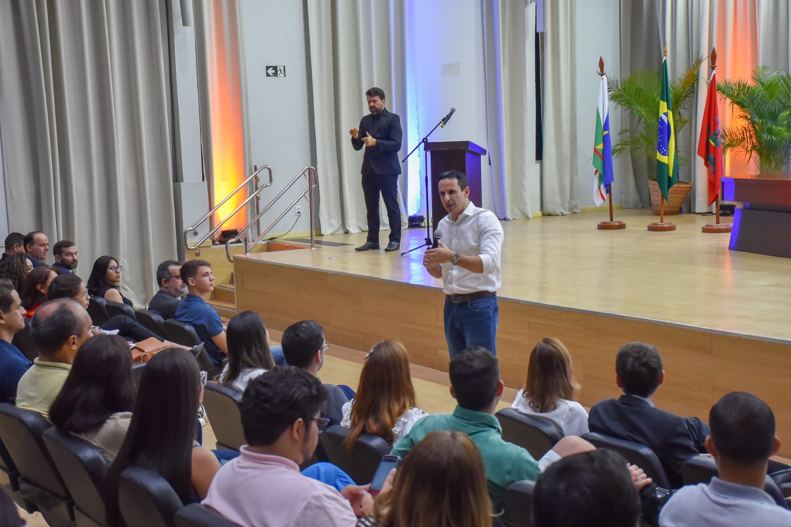  Foto colorida de uma plateia em um auditório assistindo um homem que está falando em pé. 