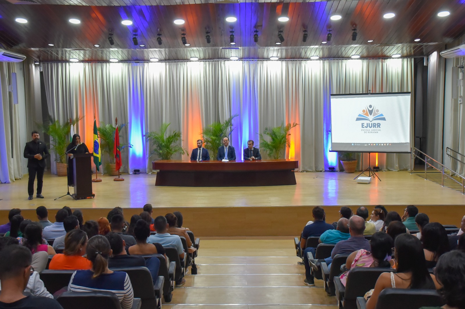 Foto colorida de um auditório lotado com três homens sentados na mesa que está no palco