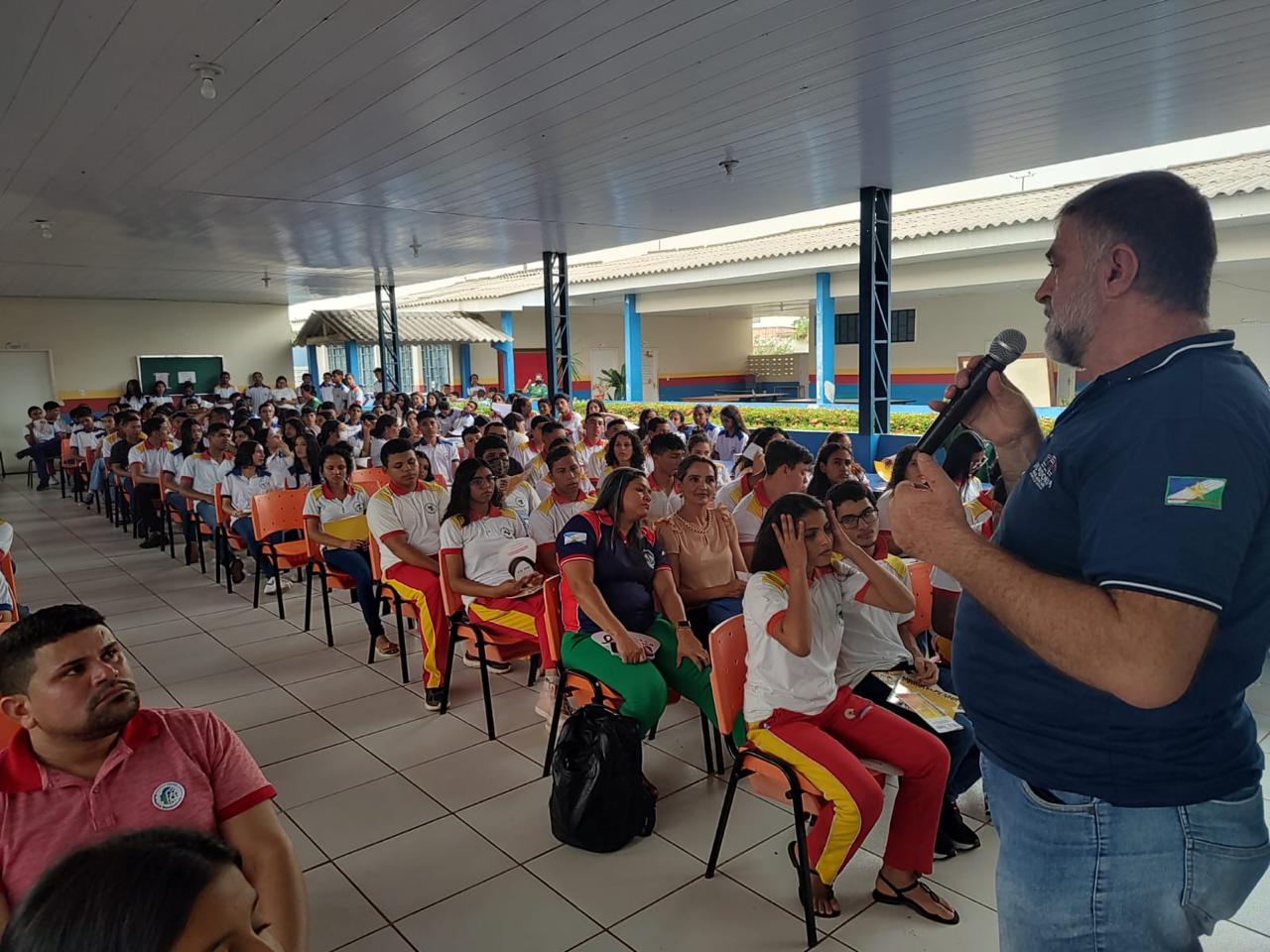  Foto colorida de  vários estudantes sentados em cadeiras olhando para o Assistente Técnico da Ouvidoria, Amadeu Triani, que está em pé falando