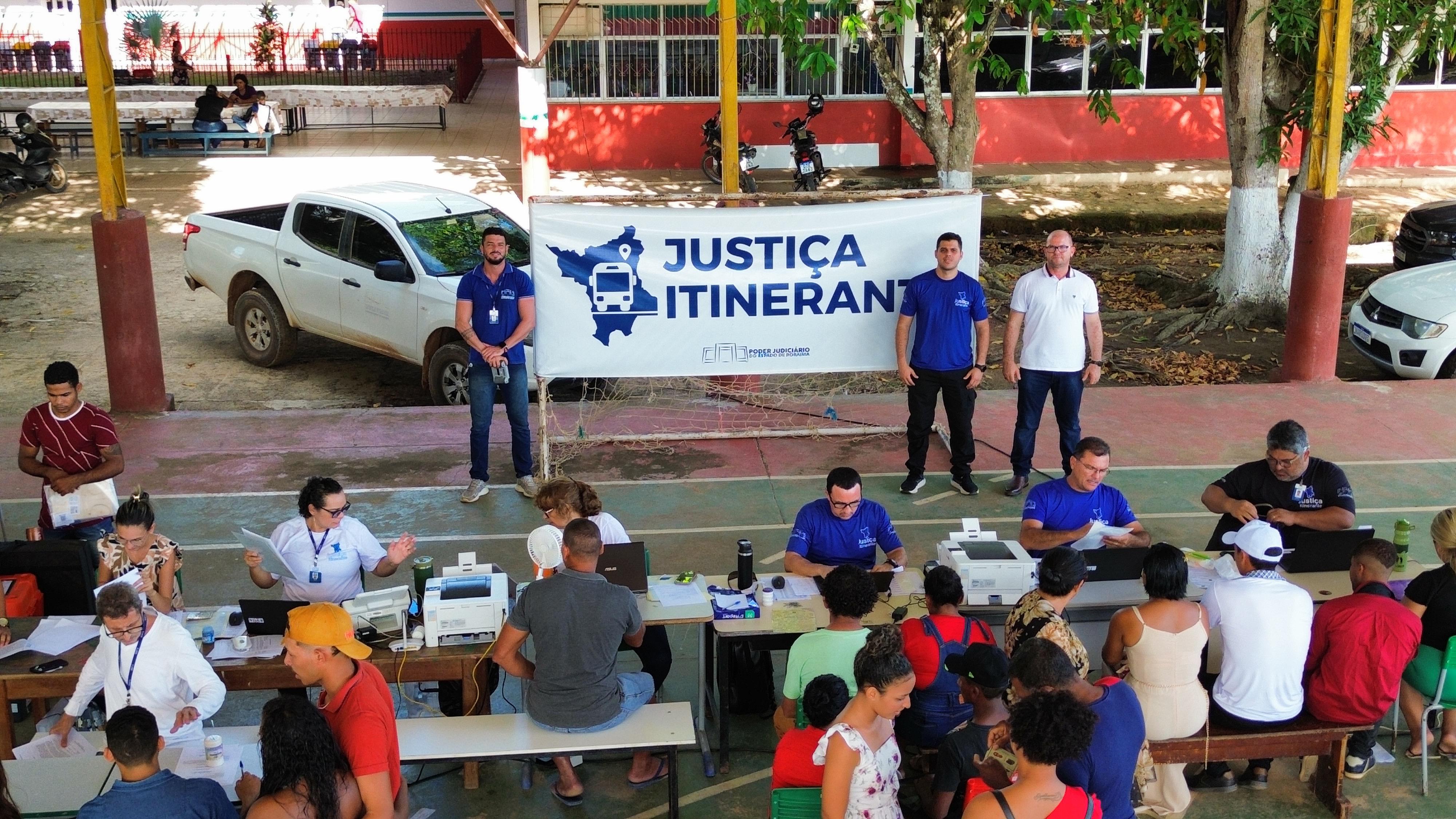 Imagem colorida mostra um grupo de servidores e servidoras do Justiça Itinerante do TJRR durante ação no interior do Estado de Roraima. Na imagem três servidores posam para fotografia ao lado do banner do Justiça itinerante do Poder Judiciário de Roraima e os outros servidores estão atendendo a população.