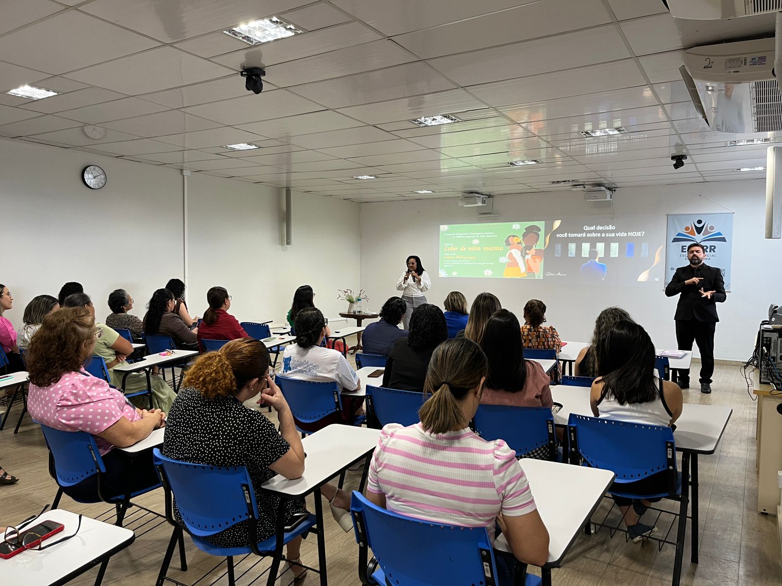 Foto colorida de mulheres uma sala de aula. Ao fundo, uma palestrante e um intérprete de libras.