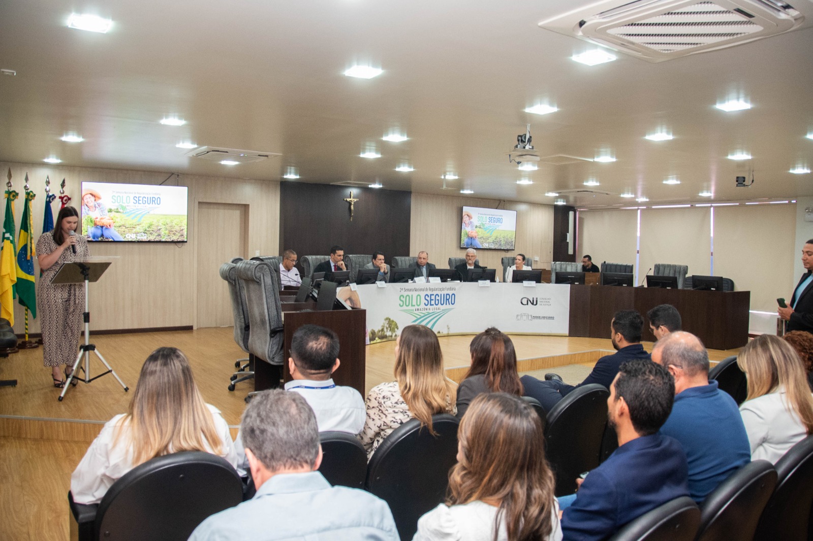 Foto colorida mostrando pessoas sentadas nas cadeiras enfileiradas do auditório do Tribunal Pleno durante a solenidade do Solo Seguro. No púlpito uma mulher ler um termo de acordo entre a Diocese e a Prefeitura. Mais a direita na imagem a mesa de autoridades. Abaixo na imagem o texto: "SOLO SEGURO - Regularização Fundiária avança em Roraima com doação de terras e entrega de títulos" .