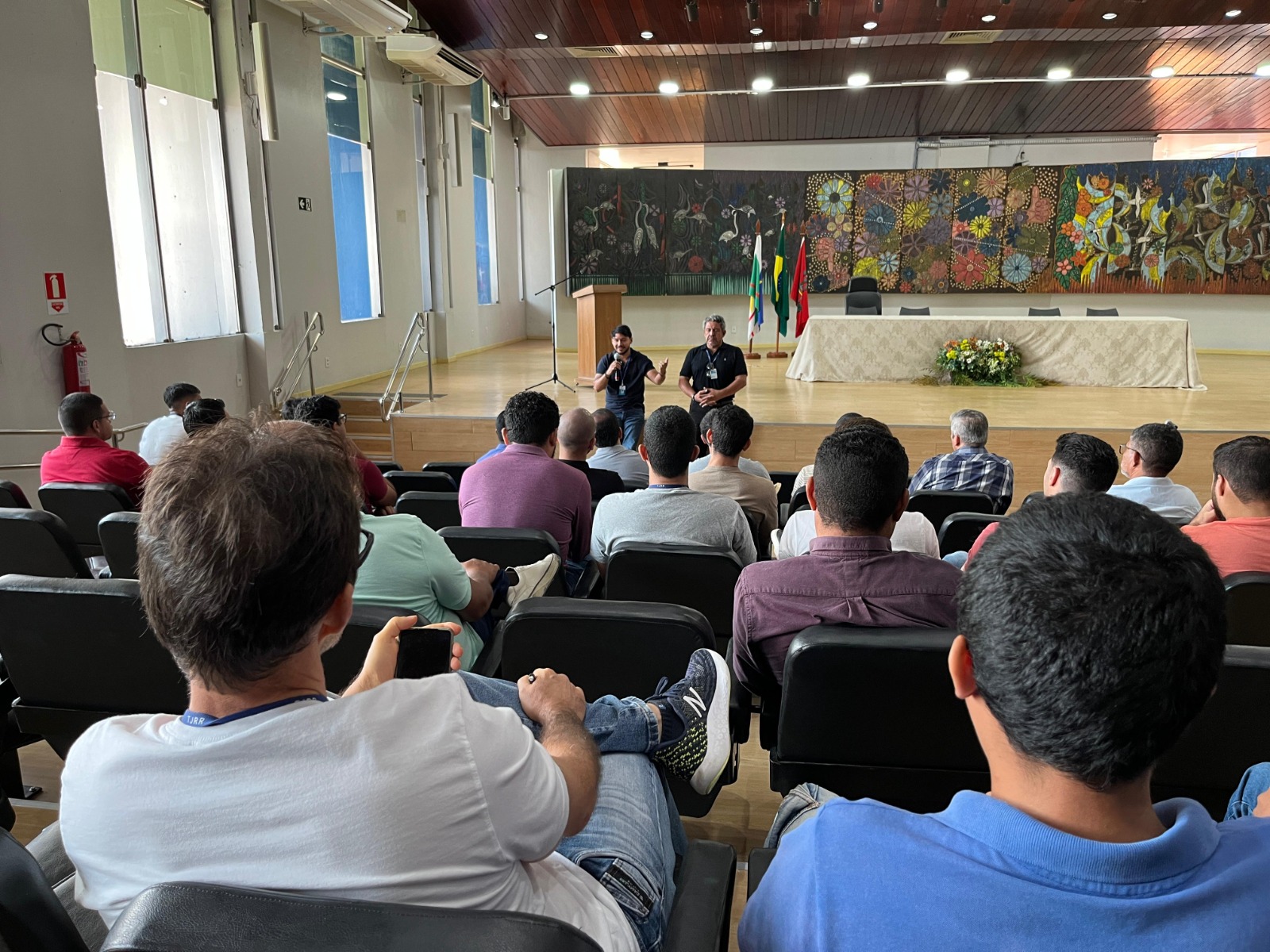 Foto Colorida mostra de frente mais a fundo o servidor da Ouvidoria-Geral do Tribunal de Justiça de Roraima, Orib Ziedson, em pé palestrando para homens sentados nas cadeiras do auditório que estão em destaque na imagem.