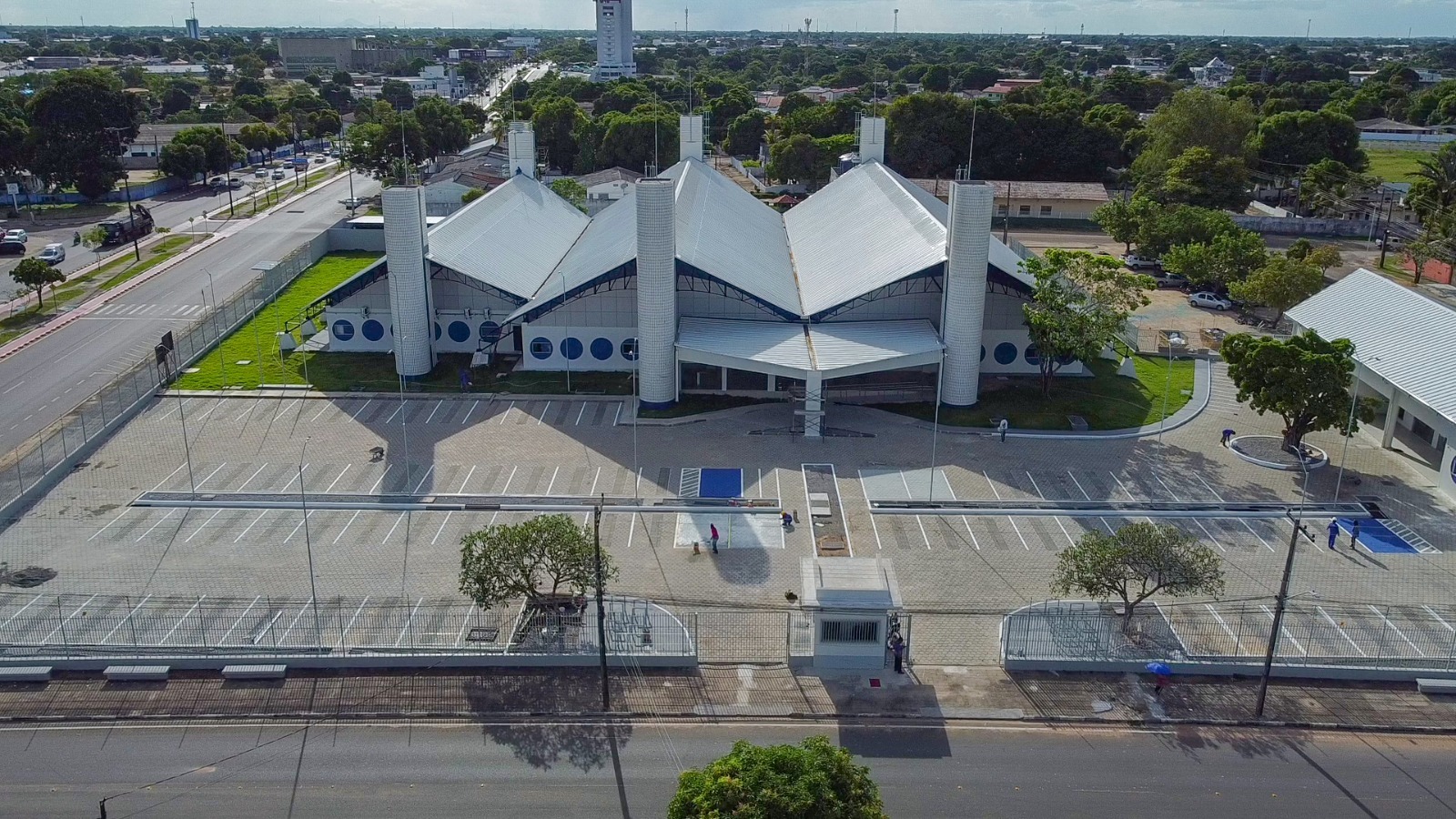 A imagem exibe o prédio do Fórum da Cidadania, com arquitetura moderna, formado por estruturas triangulares no telhado e pilares brancos. Ele está situado em uma área espaçosa, com um estacionamento amplo e arborizado ao redor. A via adjacente é larga e bem organizada, refletindo um ambiente urbano planejado.