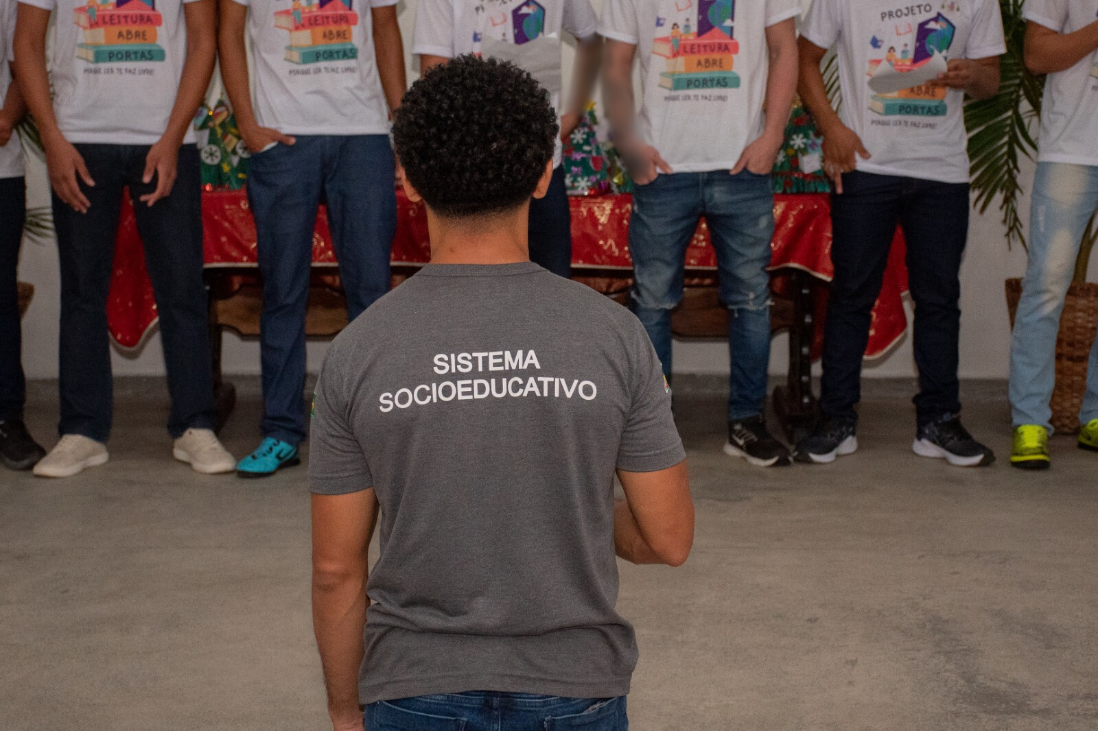 foto colorida de uma pessoa de costas com blusa cinza escrita "Sistema SocioEducativo" e sua frente tem tem jovens aparecendo da cintura para baixo.