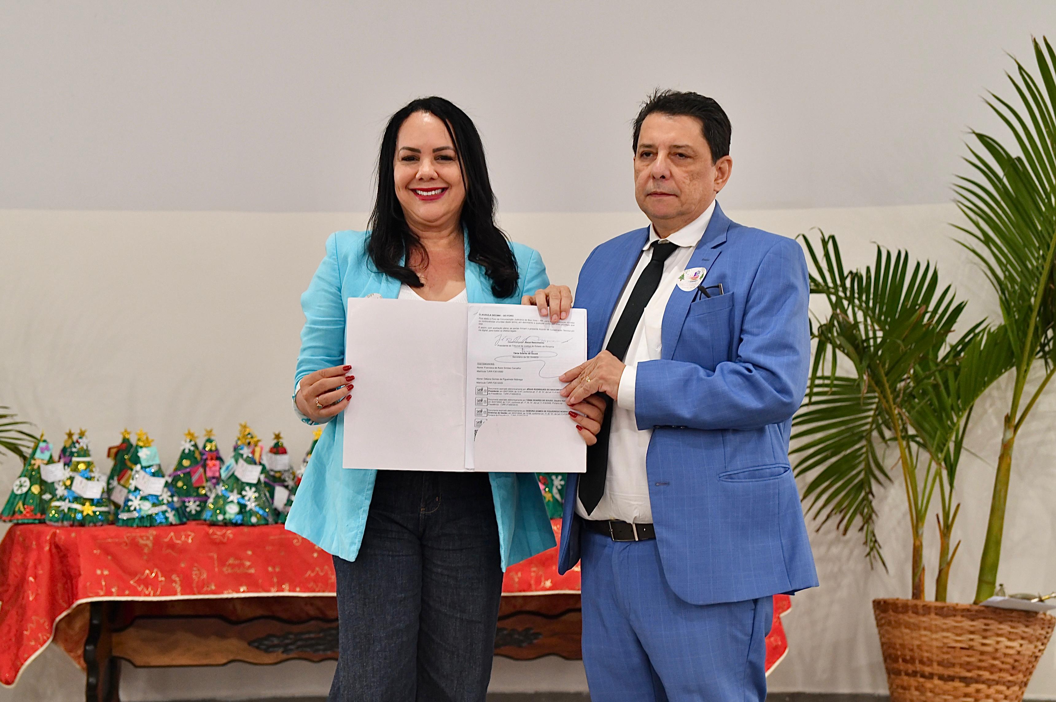 foto colorida do presidente do TJRR, Jésus Nascimento, segurando junto à Secretaria do Setrabes, Tânia Vasconcelos o termo de cooperação.