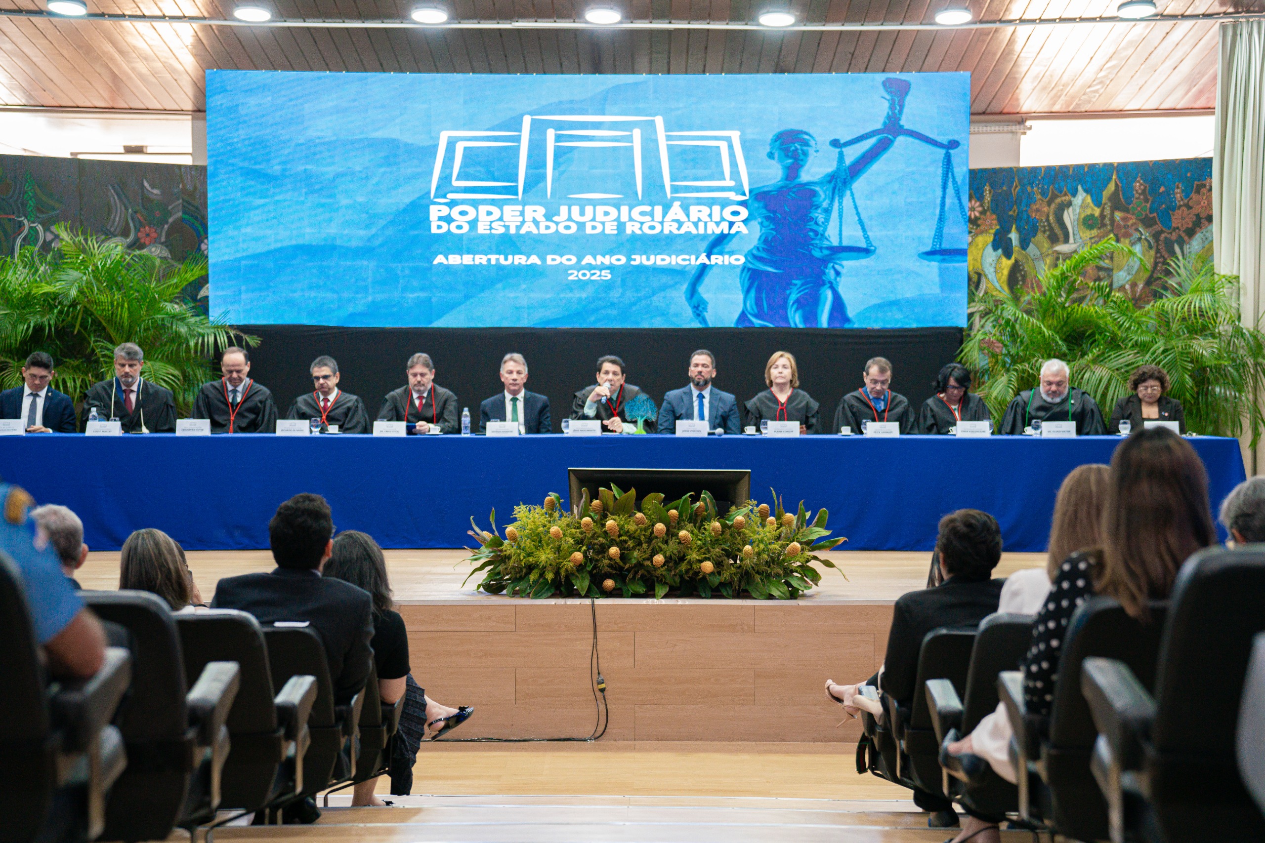 foto colorida mostra a solenidade de abertura do Ano Judiciário 2025 do Tribunal de Justiça de Roraima (TJRR). No palco, uma mesa com treze autoridades veste trajes formais e togas. Da esquerda para a direita: o presidente da Associação de Magistrados do Estado de Roraima, Marcelo Oliveira; o juiz convocado, Luiz Fernando Mallet; o diretor da Escola Judicial de Roraima, desembargador Cristóvão Súter; o vice-presidente do Tribunal de Justiça de Roraima, desembargador Ricardo Oliveira; o procurador-geral de Justiça do Ministério Público de Roraima, Fábio Stica; o governador do Estado de Roraima, Antonio Denarium; o presidente do Tribunal de Justiça do Estado de Roraima, desembargador Jésus Nascimento; o vice-presidente da Assembleia Legislativa do Estado de Roraima, deputado Jorge Everton; a presidente do TRE-Roraima, desembargadora Elaine Bianchi; o ouvidor-geral de Justiça, desembargador Erick Linhares; a corregedora do TRE, desembargadora Tânia Vasconcelos; o defensor público-geral do Estado de Roraima, Dr. Oleno Matos; e a vice-presidente da Ordem dos Advogados do Brasil – Seccional Roraima, Dra. Natália Leitão Costa. Ao fundo, um telão exibe o logotipo do TJRR e a inscrição "Abertura do Ano Judiciário 2025".