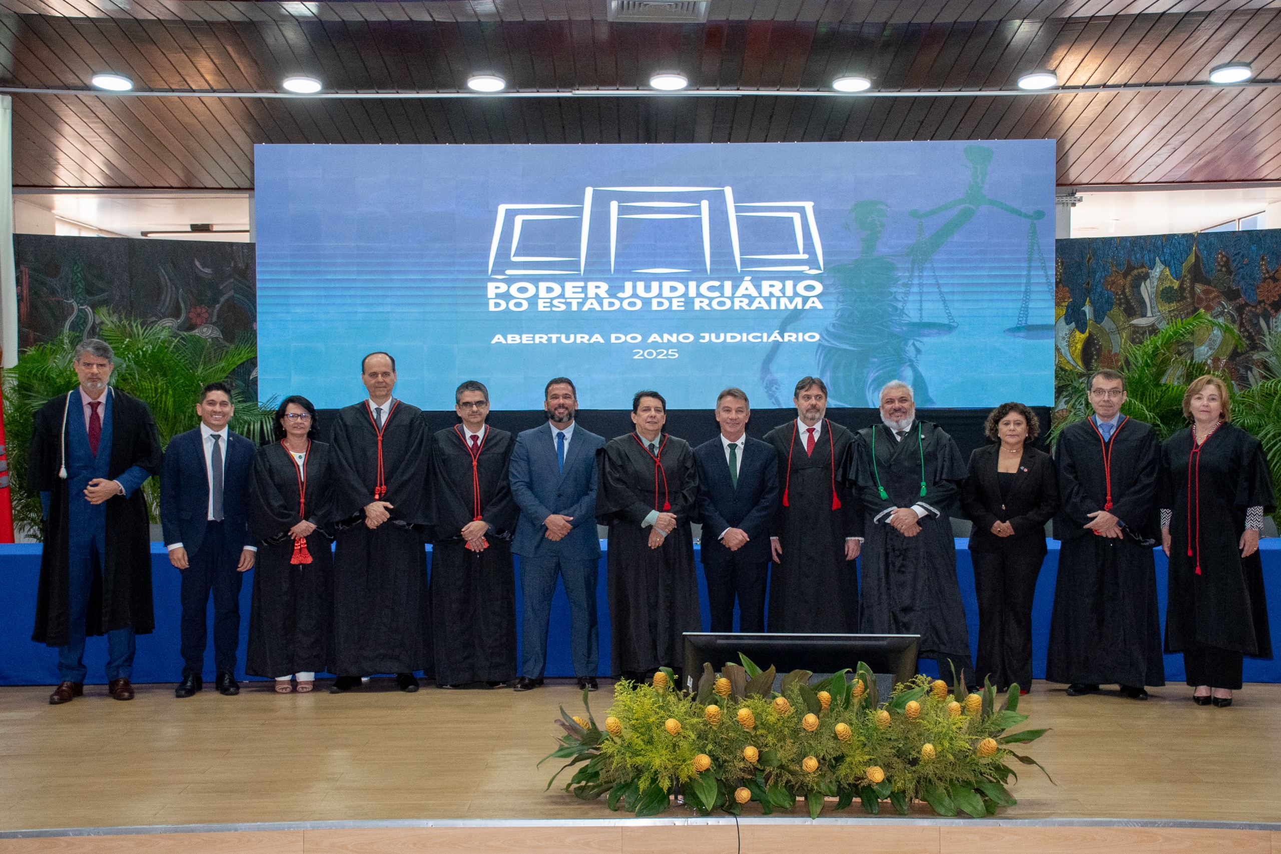 foto colorida mostra treze autoridades posando em pé para uma foto oficial durante a abertura do Ano Judiciário 2025 do Tribunal de Justiça de Roraima. Da esquerda para a dirita: o juiz convocado, Luiz Fernando Mallet; o presidente da Associação de Magistrados do Estado de Roraima, Marcelo Oliveira; a corregedora do TRE, desembargadora Tânia Vasconcelos; o diretor da Escola Judicial de Roraima, desembargador Cristóvão Súter; o vice-presidente do Tribunal de Justiça de Roraima, desembargador Ricardo Oliveira; o vice-presidente da Assembleia Legislativa do Estado de Roraima, deputado Jorge Everton; o vice-presidente da Assembleia Legislativa do Estado de Roraima, deputado Jorge Everton; o presidente do Tribunal de Justiça do Estado de Roraima, desembargador Jésus Nascimento; o governador do Estado de Roraima, Antônio Denarium; o procurador-geral de Justiça do Ministério Público de Roraima, Fábio Stica; o defensor público-geral do Estado de Roraima, Dr. Oleno Matos; a vice-presidente da Ordem dos Advogados do Brasil – Seccional Roraima, Dra. Natália Leitão Costa; o ouvidor-geral de Justiça, desembargador Erick Linhares; e a presidente do TRE-Roraima, desembargadora Elaine Bianchi. Eles estão no palco, vestidos com trajes formais e togas, à frente de um telão que exibe o logotipo do TJRR e a inscrição "Abertura do Ano Judiciário 2025". 