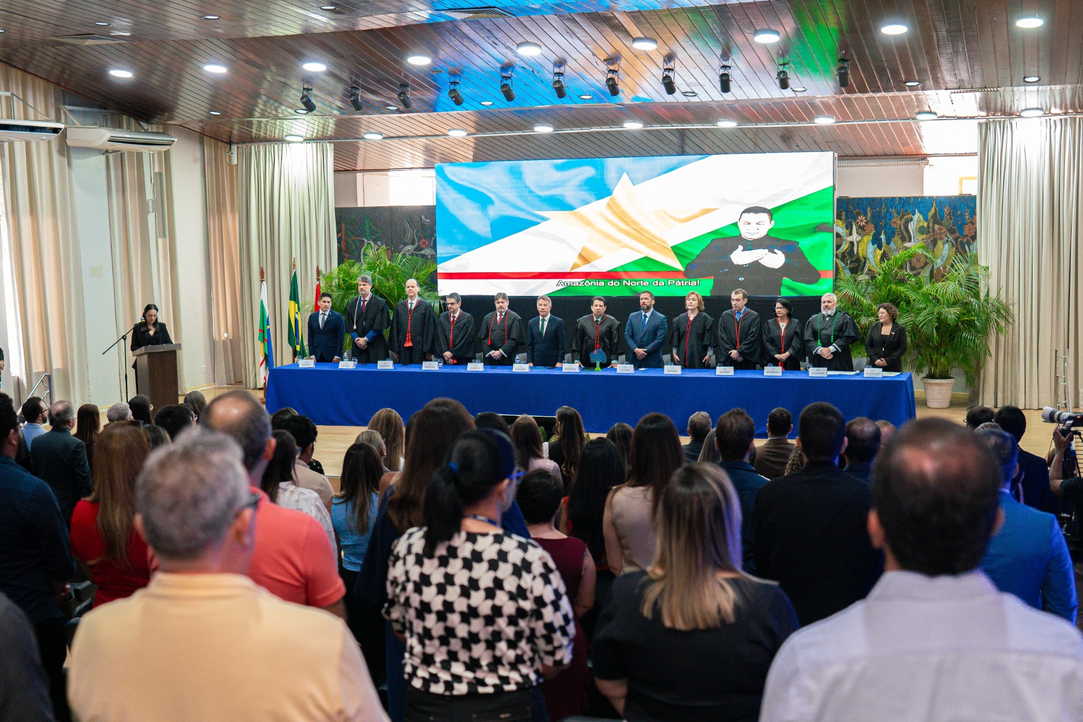 foto colorida mostra a solenidade de abertura do Ano Judiciário 2025 do Tribunal de Justiça de Roraima (TJRR). No palco, uma mesa com treze autoridades veste trajes formais e togas, enquanto uma mulher discursa em um púlpito. Da esquerda para a direita: o presidente da Associação de Magistrados do Estado de Roraima, Marcelo Oliveira; o juiz convocado, Luiz Fernando Mallet; o diretor da Escola Judicial de Roraima, desembargador Cristóvão Súter; o vice-presidente do Tribunal de Justiça de Roraima, desembargador Ricardo Oliveira; o procurador-geral de Justiça do Ministério Público de Roraima, Fábio Stica; o governador do Estado de Roraima, Antônio Denarium; o presidente do Tribunal de Justiça do Estado de Roraima, desembargador Jésus Nascimento; o vice-presidente da Assembleia Legislativa do Estado de Roraima, deputado Jorge Everton; a presidente do TRE-Roraima, desembargadora Elaine Bianchi; o ouvidor-geral de Justiça, desembargador Erick Linhares; a corregedora do TRE, desembargadora Tânia Vasconcelos; o defensor público-geral do Estado de Roraima, Dr. Oleno Matos; e a vice-presidente da Ordem dos Advogados do Brasil – Seccional Roraima, Dra. Natália Leitão Costa. Ao fundo, um telão exibe o logotipo do TJRR e a inscrição "Abertura do Ano Judiciário 2025".