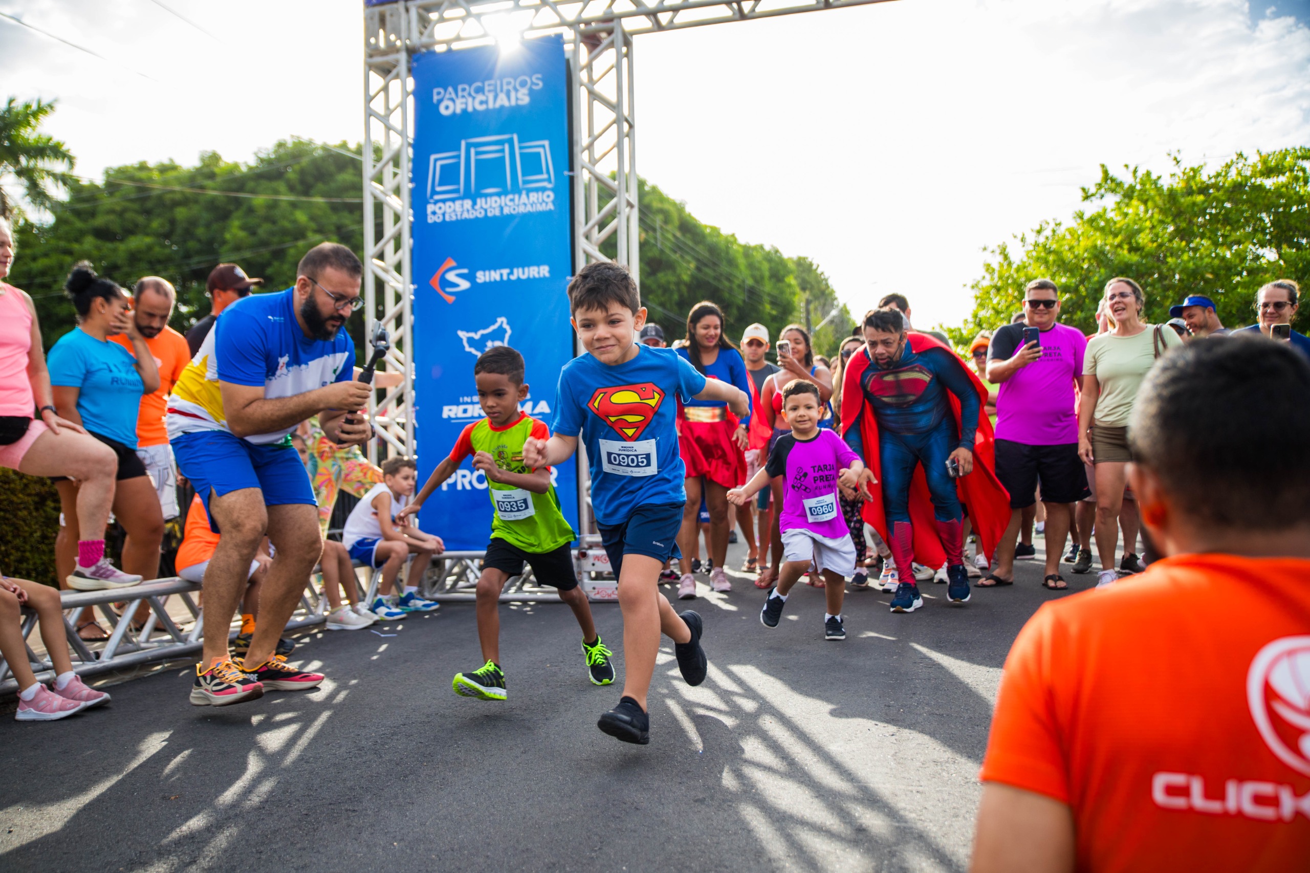  foto colorida em ambiente externo arborizado de crianças partindo da largada, a frente tem uma criança correndo com blusa azul com símbolo do super-homem.