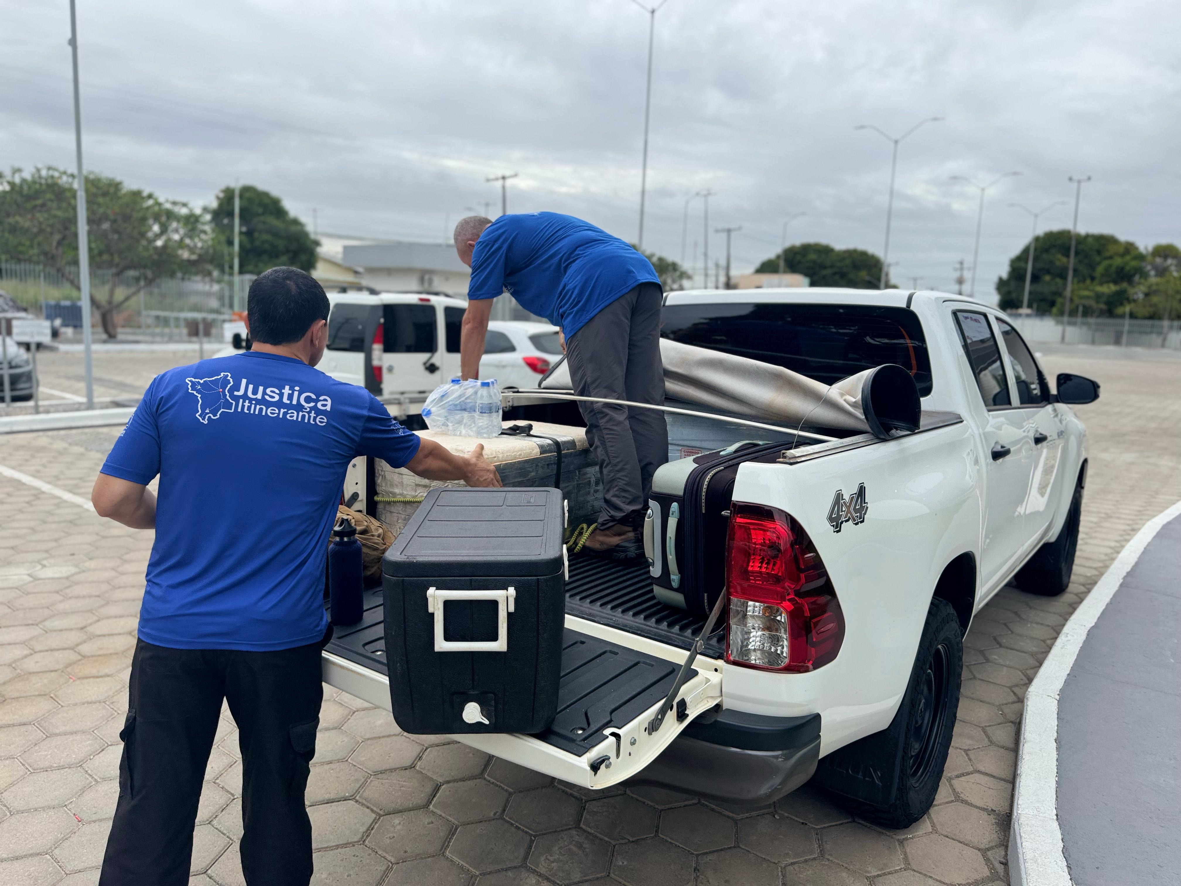 Foto colorida mostrando duas pessoas colocando uma caixa de cooler, usado para deixar bebidas geladas com o auxilio de gelo, dentro da carroceria de um carro. As duas pessoas usam uma camisa azul escrito Justiça Itinerante.