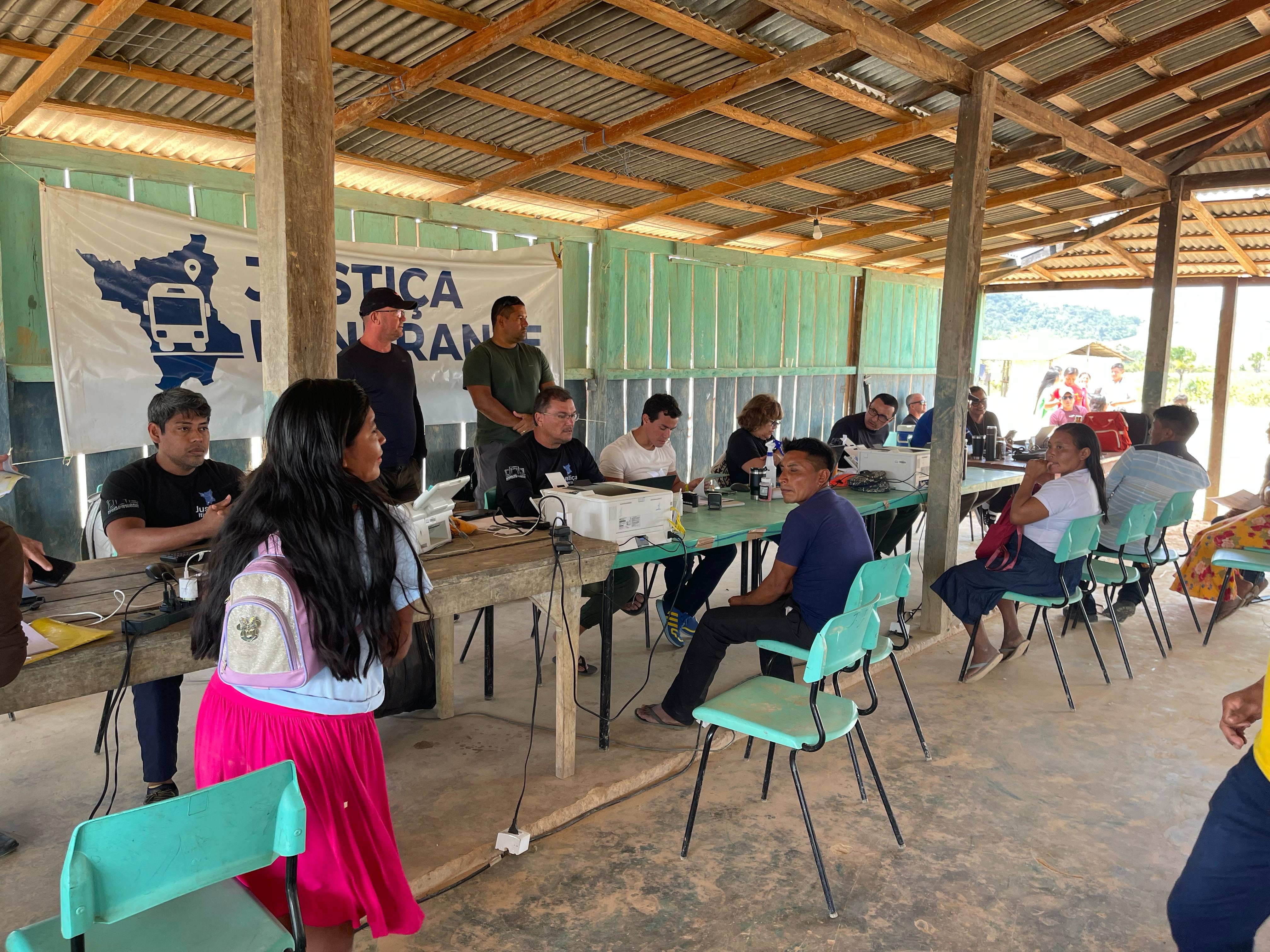 Foto colorida mostrando pessoas esperando serem atendidas pela justiça itinerante. Na imagem, servidores da justiça itnerante estão sentados atras de mesas realizando atendimentos, enquanto, do outro lado, a população espera atendimento Foto colorida mostrando pessoas esperando serem atendidas pela justiça itinerante. Na imagem, servidores da justiça itnerante estão sentados atras de mesas realizando atendimentos, enquanto, do outro lado, a população espera atendimento