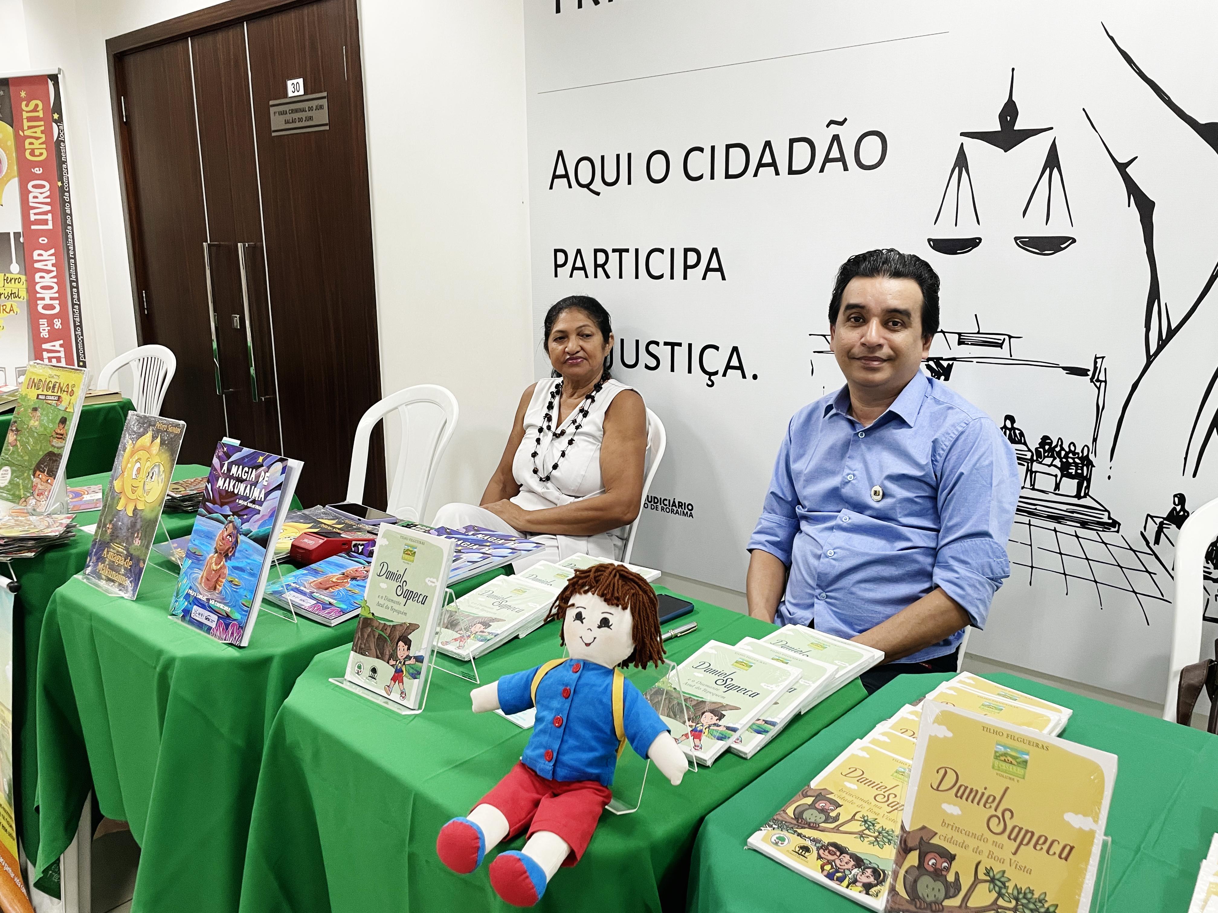 foto colorida com Clotilho Filgueiras e Pétira Santos, sentados e uma mesa à frente cheia de suas obras expostas, posando para fotografia. foto colorida com Clotilho Filgueiras e Pétira Santos, sentados e uma mesa à frente cheia de suas obras expostas, posando para fotografia.