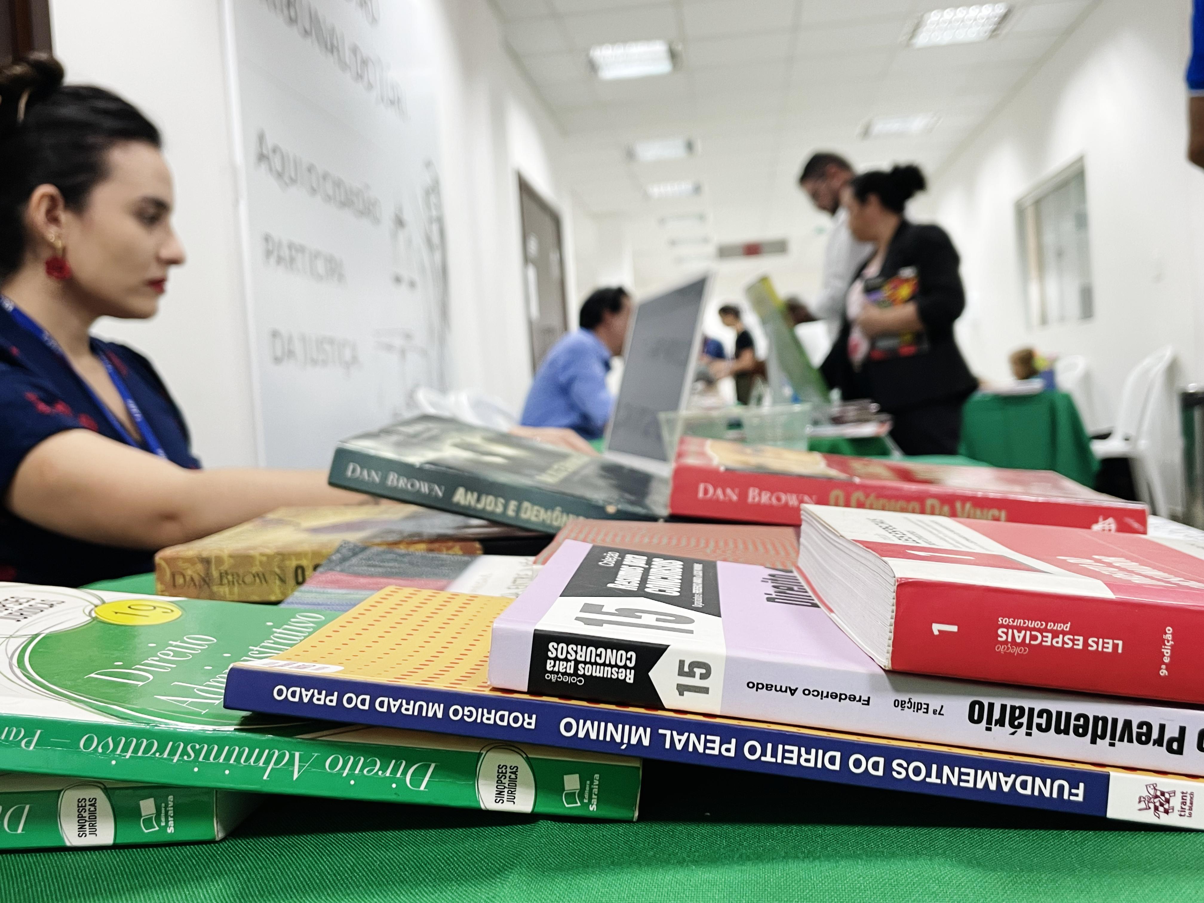 foto colorida dando enfoque em cinco livros, ao fundo tem duas pessoas realizando a compra de um livro foto colorida dando enfoque em cinco livros, ao fundo tem duas pessoas realizando a compra de um livro