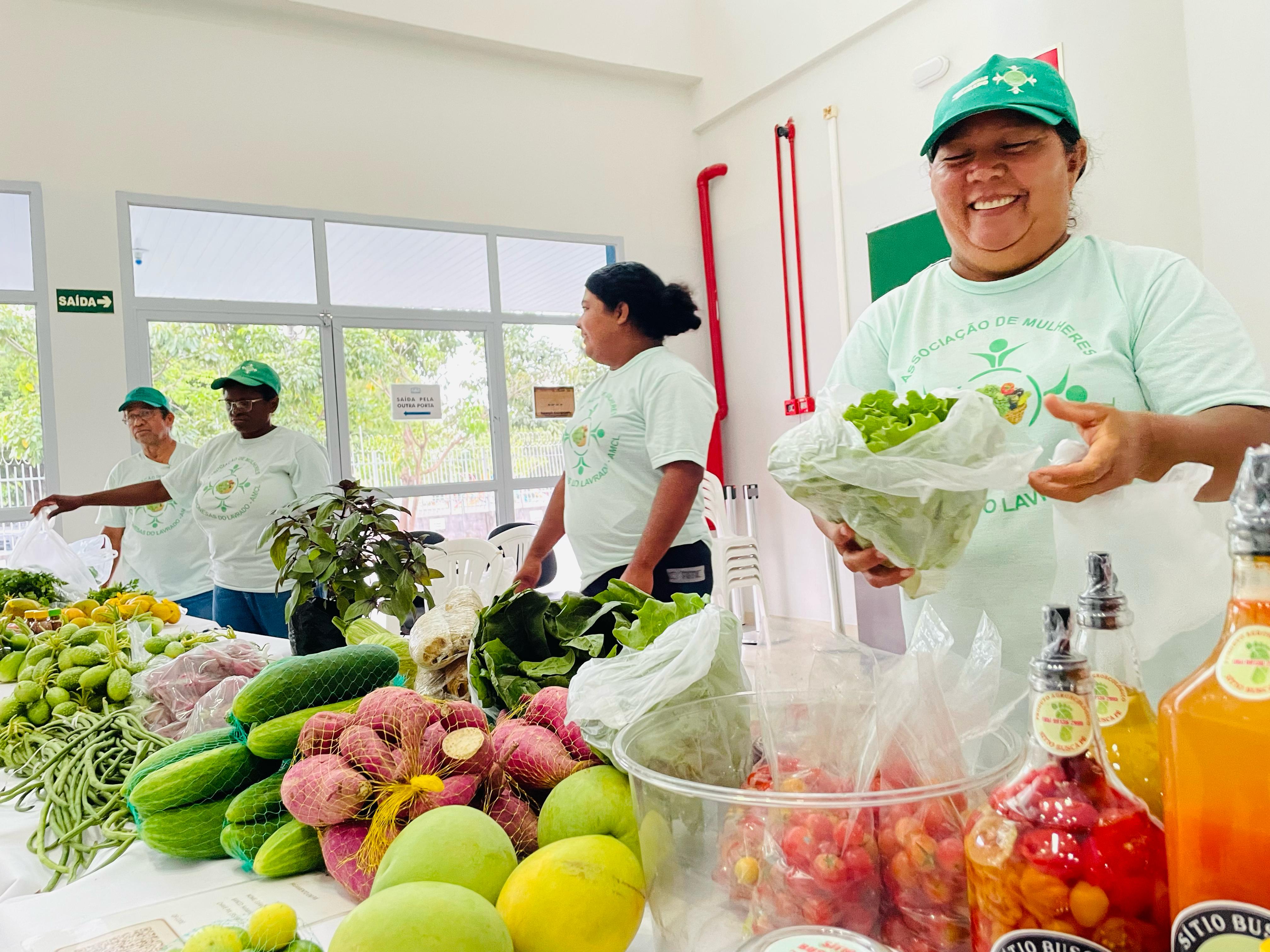 Foto colorida mostrando uma mullher segurando uma sacola com alguimas verduras dentro. Abaixo dela, uma mesa cheia de verduras e demais produtos Foto colorida mostrando uma mullher segurando uma sacola com alguimas verduras dentro. Abaixo dela, uma mesa cheia de verduras e demais produtos