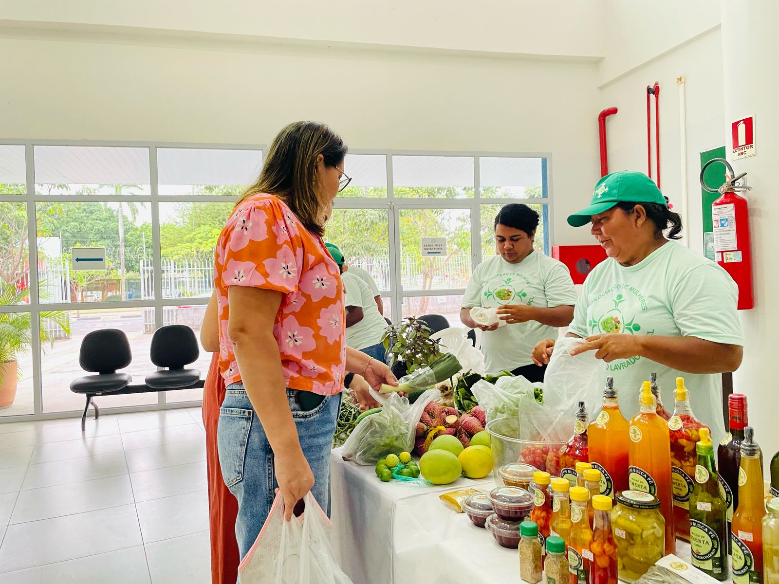 Foto colorida mostrando uma mulher vendendo alguns produtos como verduras, legumes, geleias, pimentas e entre outros. Na frente dela, duas outras mulheres estão olhando os produtos Foto colorida mostrando uma mulher vendendo alguns produtos como verduras, legumes, geleias, pimentas e entre outros. Na frente dela, duas outras mulheres estão olhando os produtos