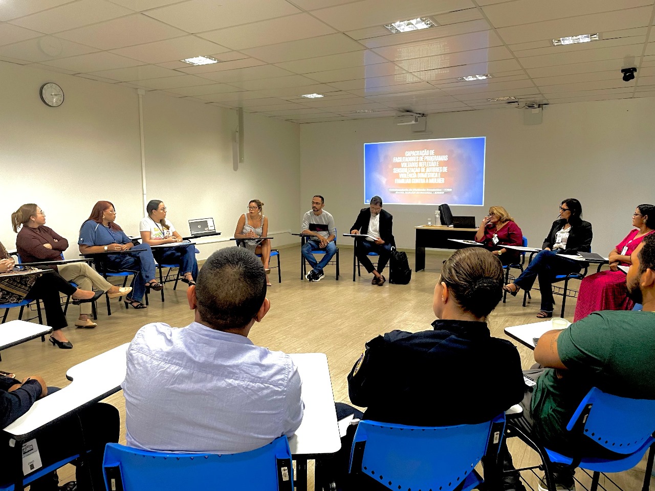Imagem colorida contém um grupo de pessoas sentadas em círculo em uma sala de reunião, participando de uma capacitação sobre atendimento a mulheres vítimas de violência, com um projetor exibindo o tema ao fundo. Imagem colorida contém um grupo de pessoas sentadas em círculo em uma sala de reunião, participando de uma capacitação sobre atendimento a mulheres vítimas de violência, com um projetor exibindo o tema ao fundo.