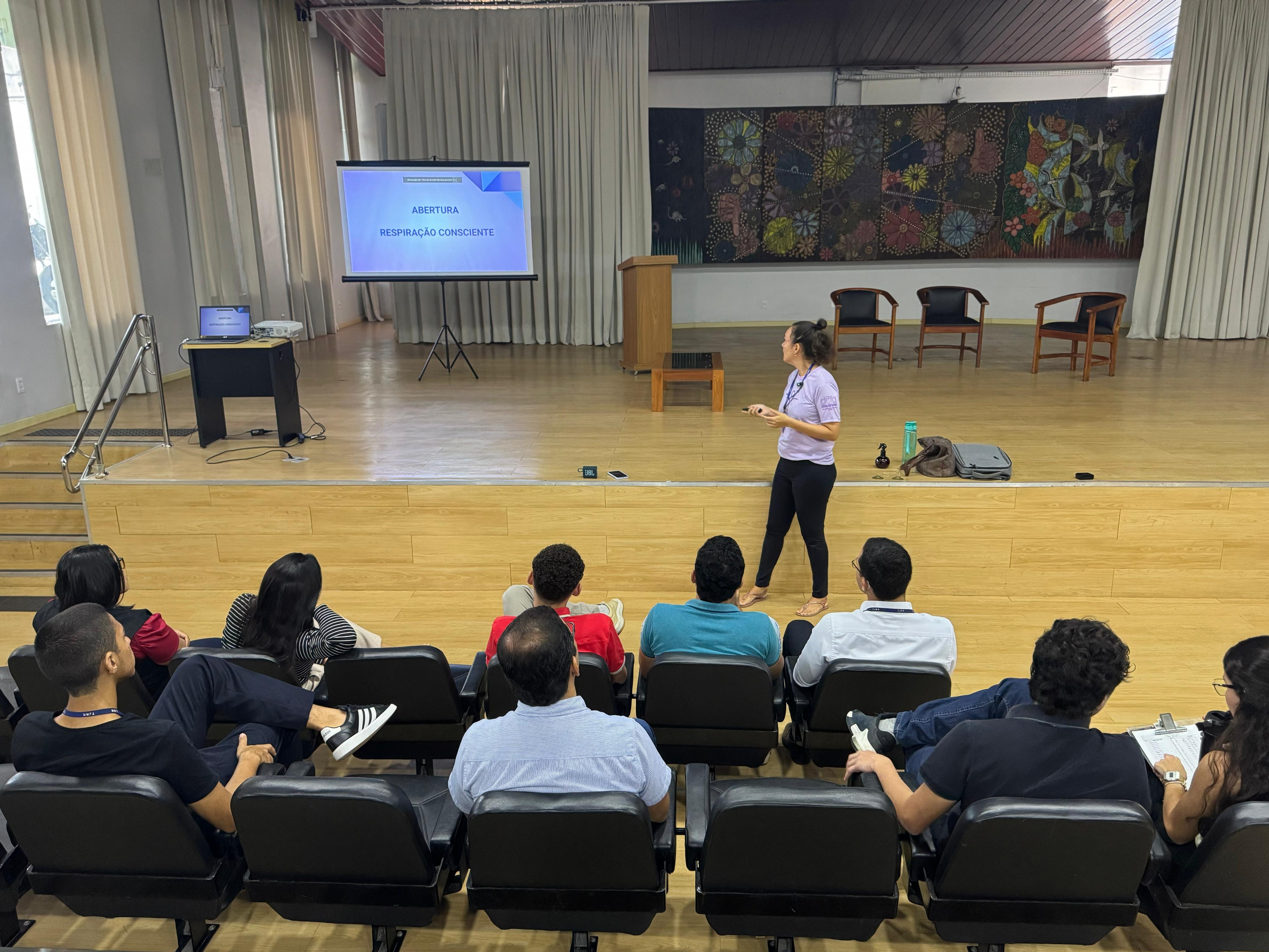 Fotografia em um auditório. Em primeiro plano, servidores do TJRR estão sentados em cadeiras pretas, voltados para o palco. No palco, uma servidora de camiseta lilás e calça preta fala ao público, posicionada de frente para a plateia. À esquerda, há um telão com o slide projetado com os dizeres “Abertura – Respiração Consciente”. Ao fundo, cortinas claras e um grande painel colorido com desenhos de flores e pássaros decoram a parede. Fotografia em um auditório. Em primeiro plano, servidores do TJRR estão sentados em cadeiras pretas, voltados para o palco. No palco, uma servidora de camiseta lilás e calça preta fala ao público, posicionada de frente para a plateia. À esquerda, há um telão com o slide projetado com os dizeres “Abertura – Respiração Consciente”. Ao fundo, cortinas claras e um grande painel colorido com desenhos de flores e pássaros decoram a parede.