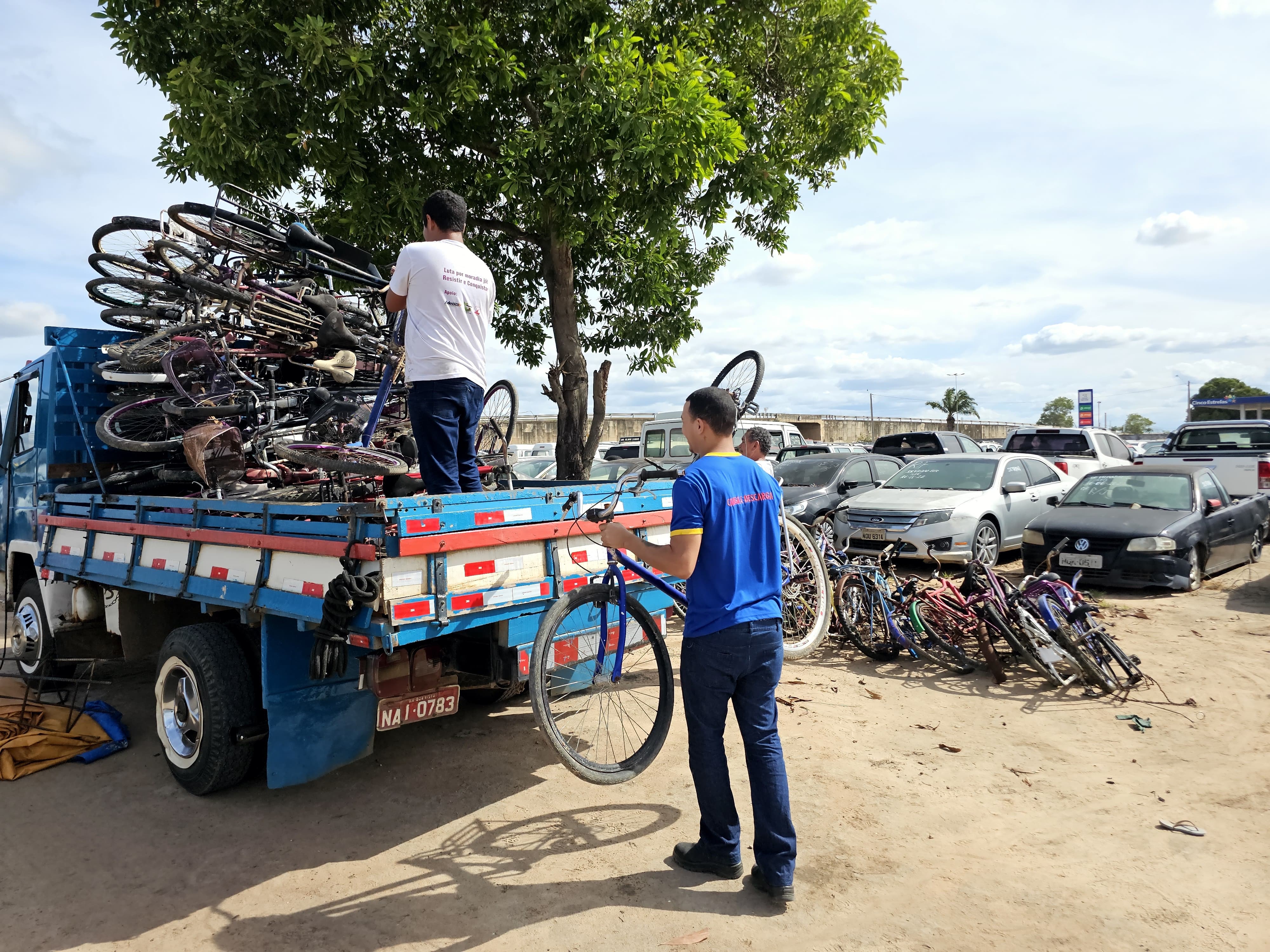 Imagem colorida contém três homens organizando as bicicletas doadas em cima de um caminhão azul. Imagem colorida contém três homens organizando as bicicletas doadas em cima de um caminhão azul.
