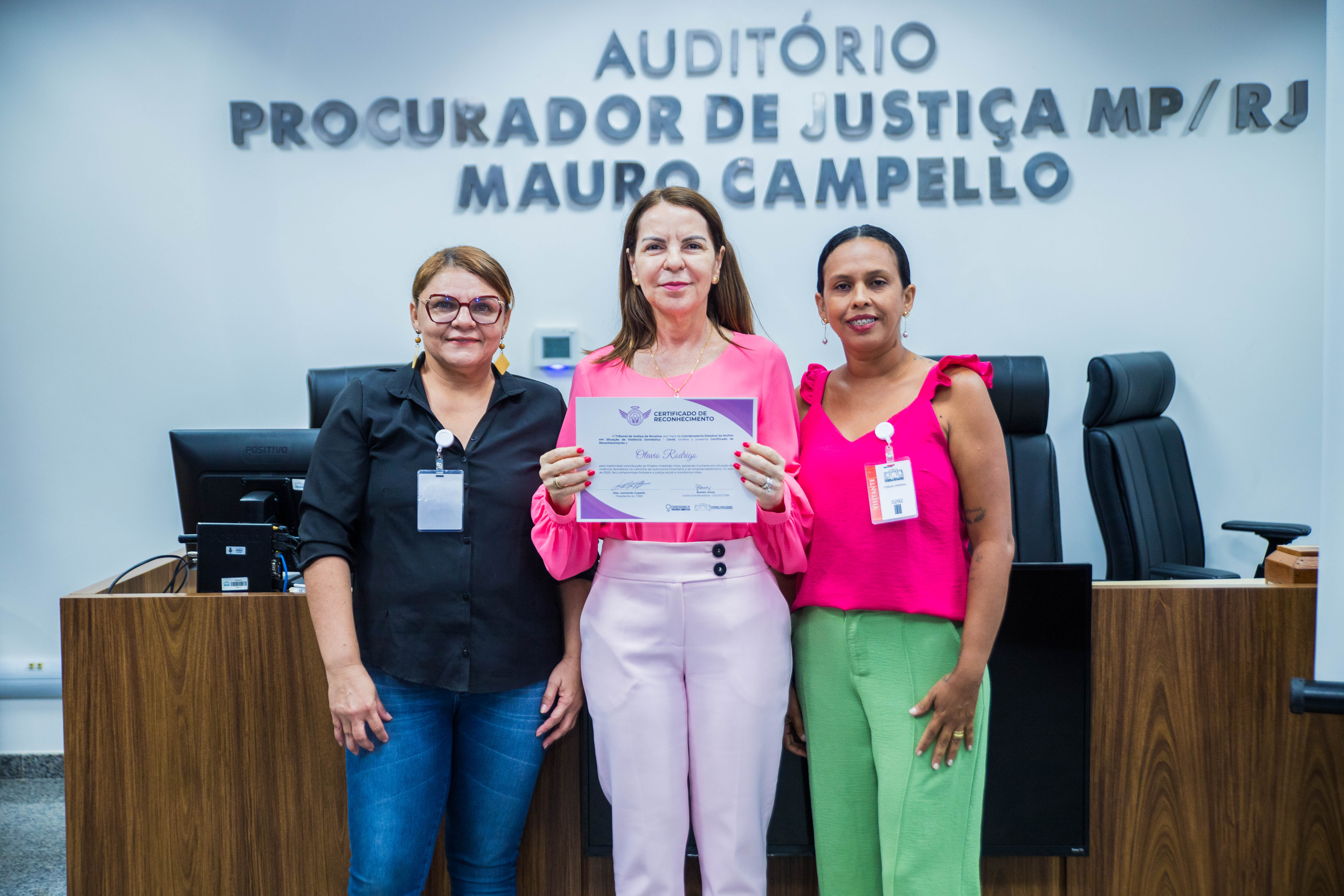 A foto mostra três mulheres adultas em pé, posando para uma foto. A mulher no centro é um pouco mais alta que as outras duas e está segurando um certificado. A foto mostra três mulheres adultas em pé, posando para uma foto. A mulher no centro é um pouco mais alta que as outras duas e está segurando um certificado.