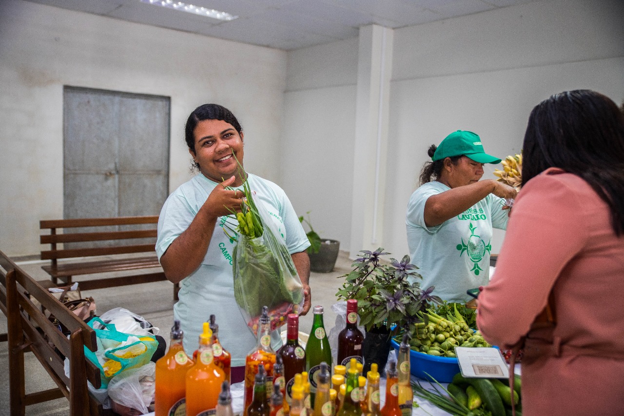 Foto colorida mostra uma feira, com duas mulheres vendendo produtos artesanais e orgânicos, como vegetais frescos, licores, azeites aromatizados e possivelmente pães ou bolos Foto colorida mostra uma feira, com duas mulheres vendendo produtos artesanais e orgânicos, como vegetais frescos, licores, azeites aromatizados e possivelmente pães ou bolos