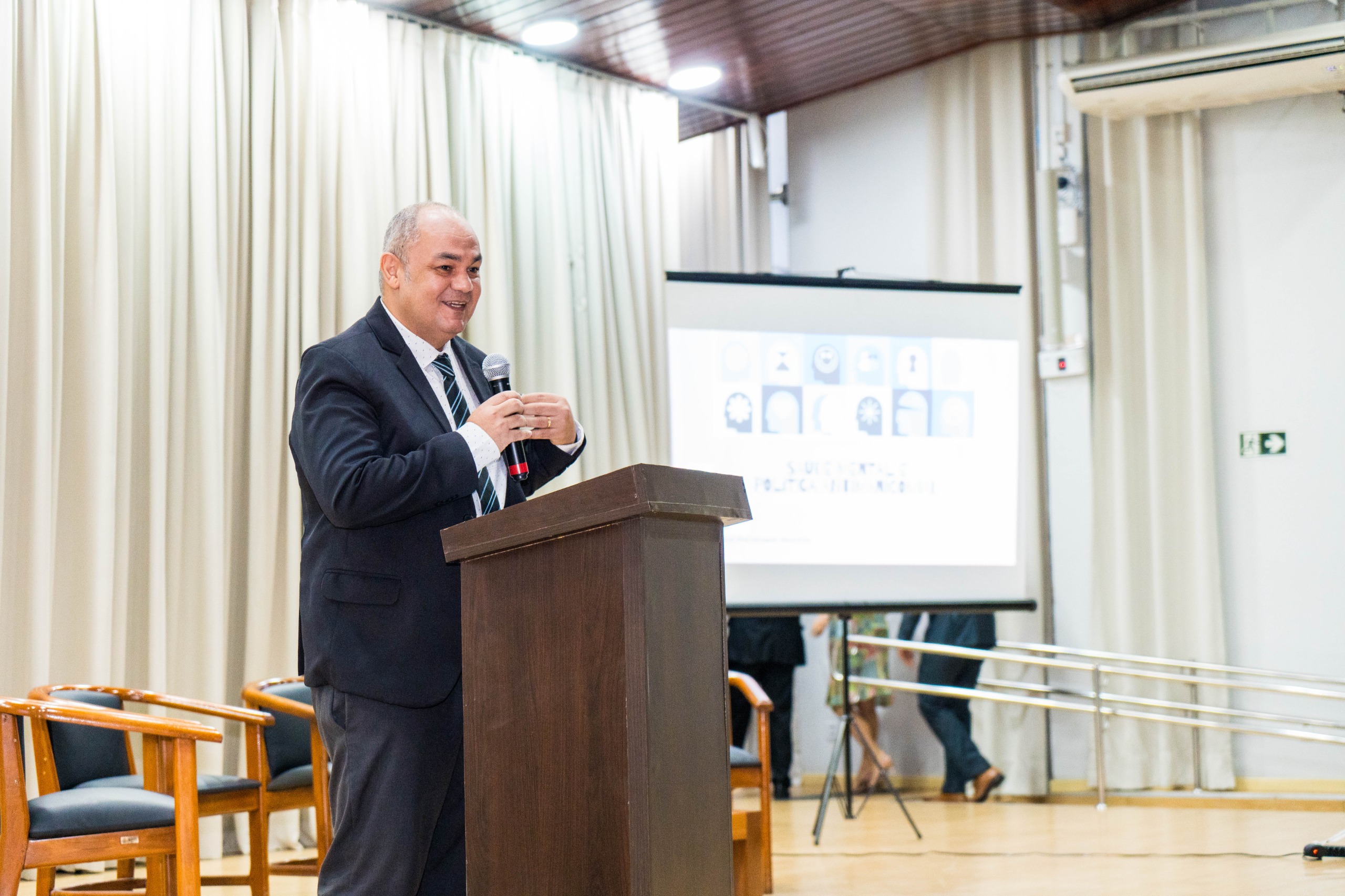 Foto colorida mostra o João Mendes Júnior, representante do Ministério da Saúde, discursando em cima do palco do auditório. Foto colorida mostra o João Mendes Júnior, representante do Ministério da Saúde, discursando em cima do palco do auditório.