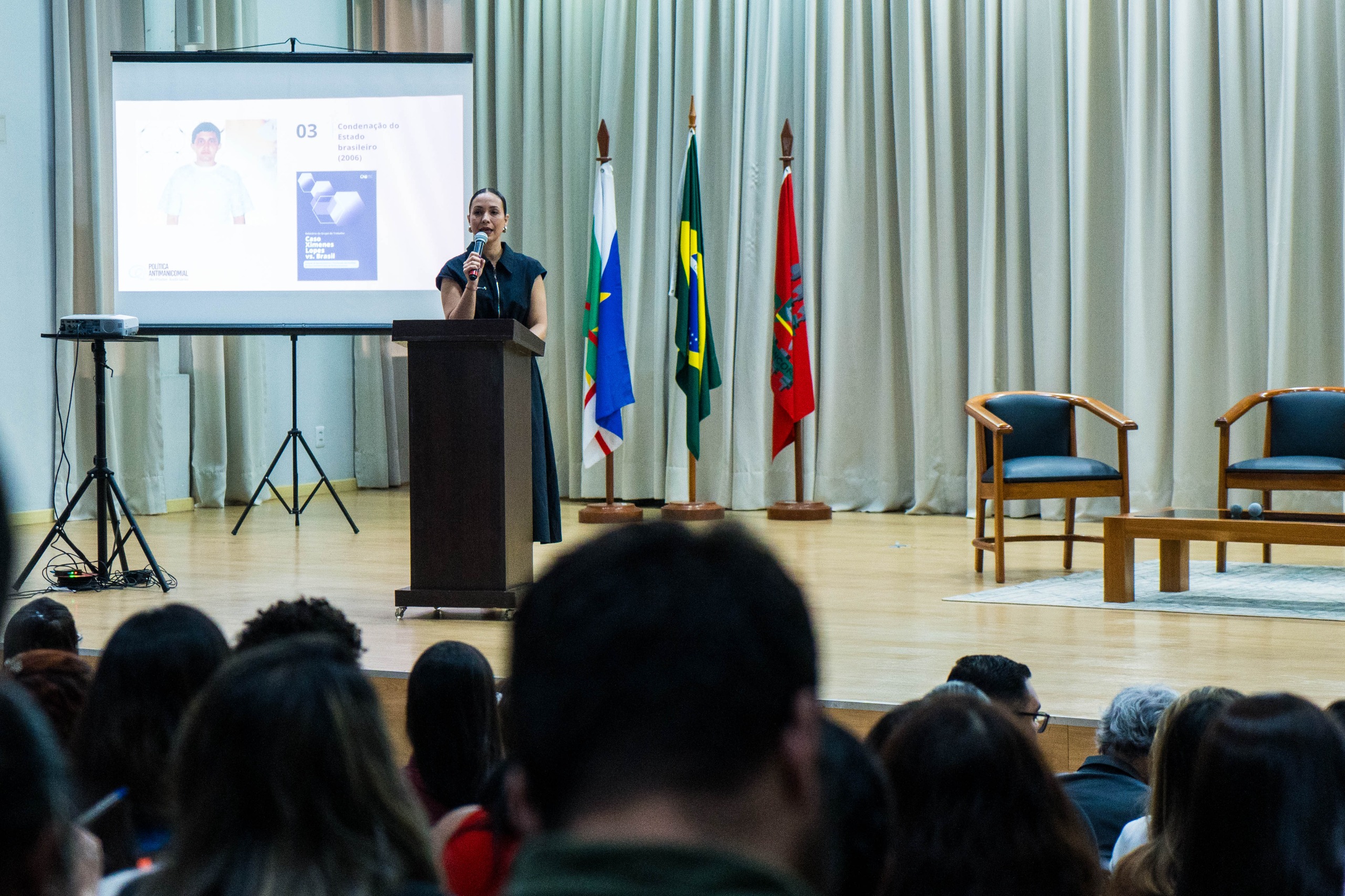 Foto colorida mostra a supervisora chefe da Seção de Apoio Institucional do Departamento de Monitoramento e Fiscalização do Sistema Carcerário e do Sistema de Execução de Medidas Socioeducativas do DMF discursando em cima do palco do auditório. Foto colorida mostra a supervisora chefe da Seção de Apoio Institucional do Departamento de Monitoramento e Fiscalização do Sistema Carcerário e do Sistema de Execução de Medidas Socioeducativas do DMF discursando em cima do palco do auditório.
